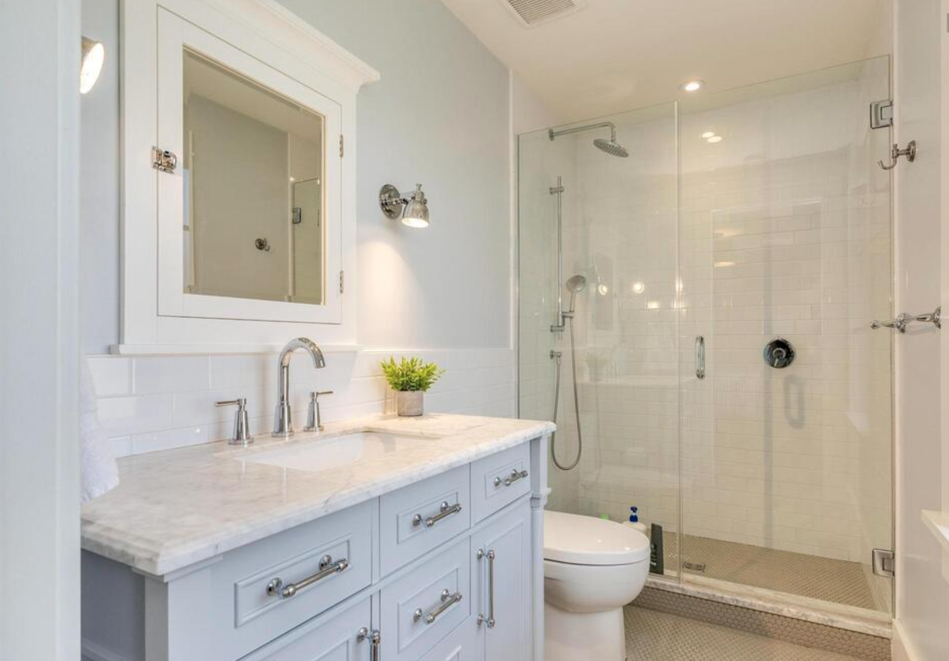A modern bathroom with a white vanity, marble countertop, a potted plant, a wall-mounted mirror, a wall-mounted light fixture, a glass-enclosed shower, a handheld showerhead, a toilet, and white tiled walls.