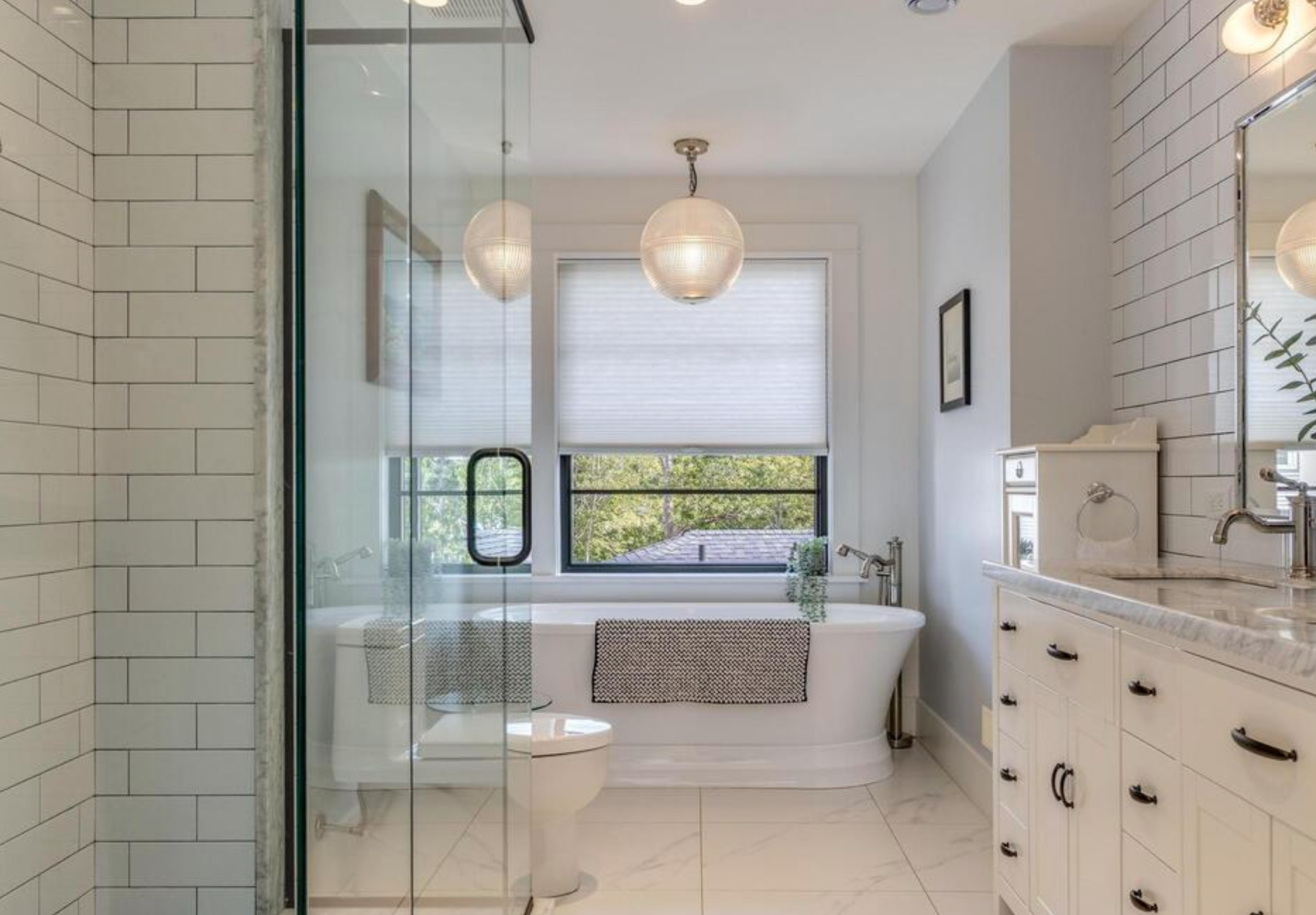 Bright bathroom with a large window, bathtub, and vanity with white cabinets and marble countertop.