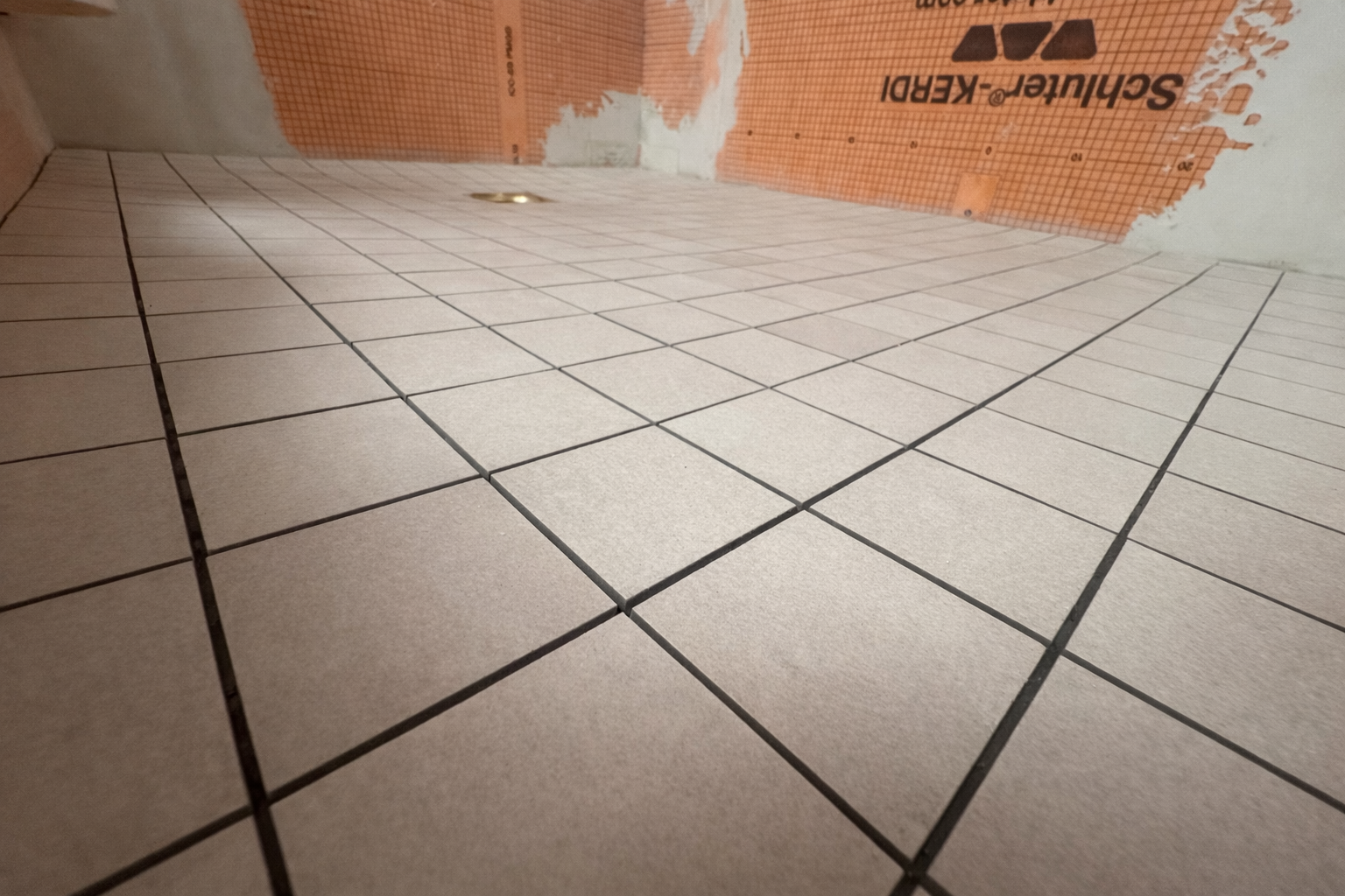 Close-up view of a tiled floor in a bathroom under construction, with white tiles and black grout lines, and a drain in the floor. There is also a partially installed orange waterproofing membrane on the wall.