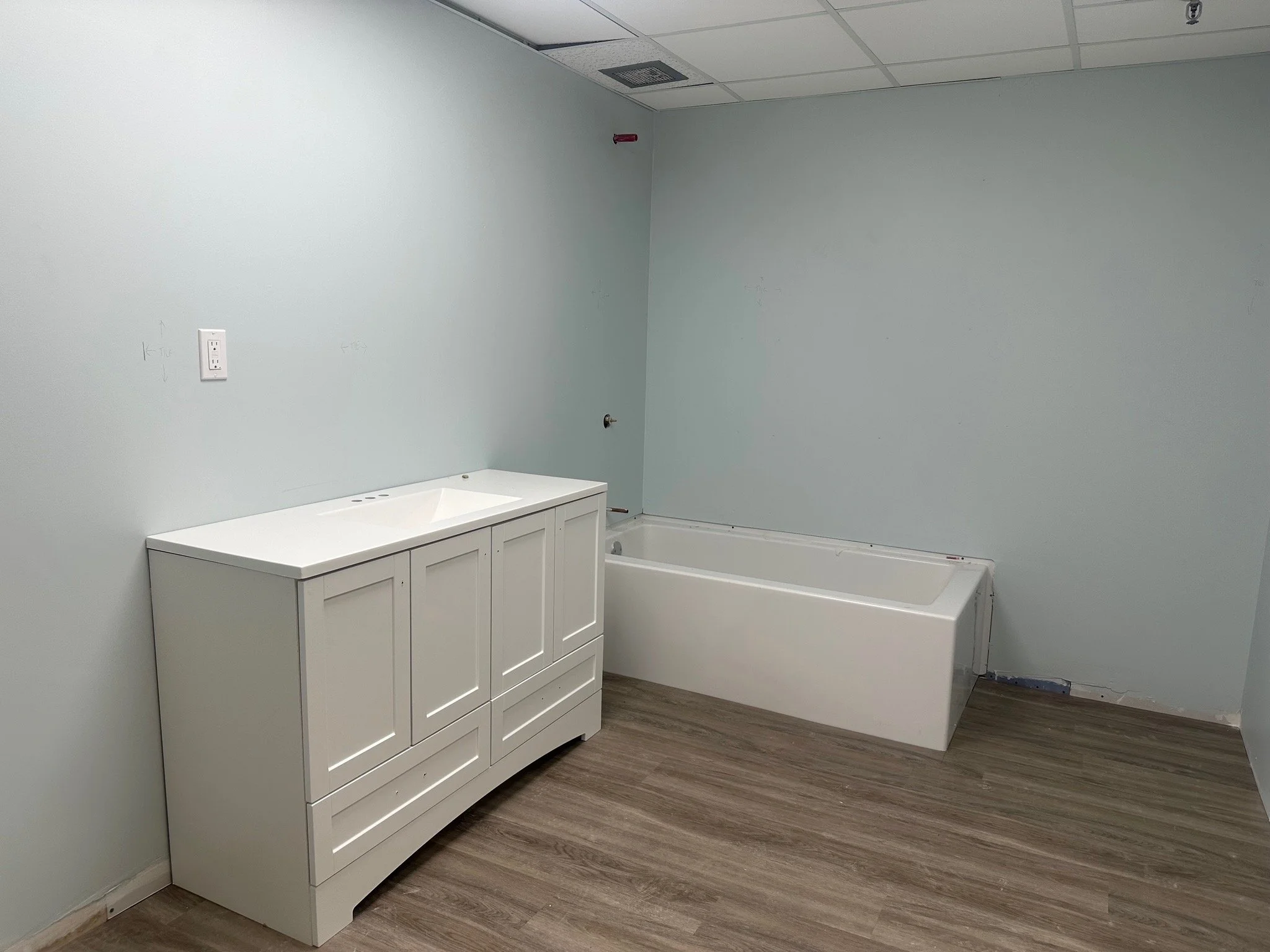Empty bathroom with a white vanity cabinet and a bathtub against light blue walls with some markings, and a wood-like laminate flooring.