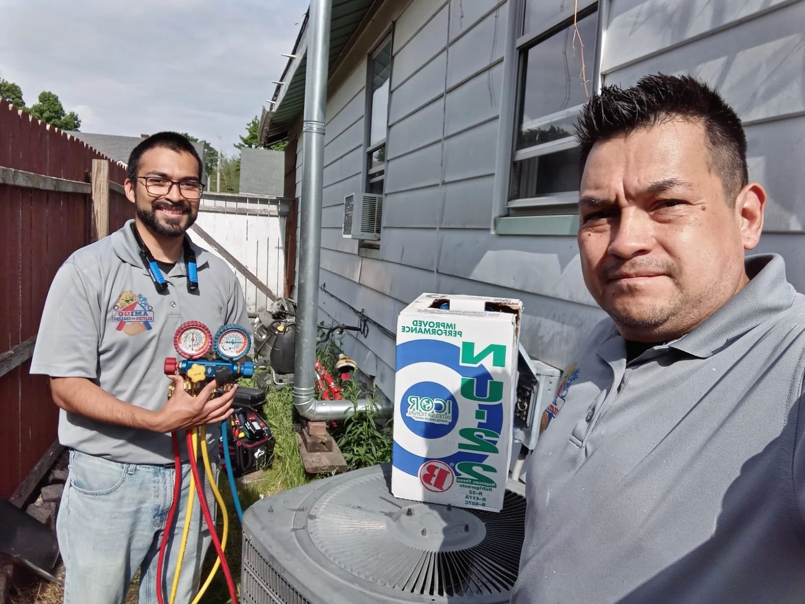 Two men working on an air conditioning unit outside a house. One man is holding HVAC tools, the other is taking a selfie.
