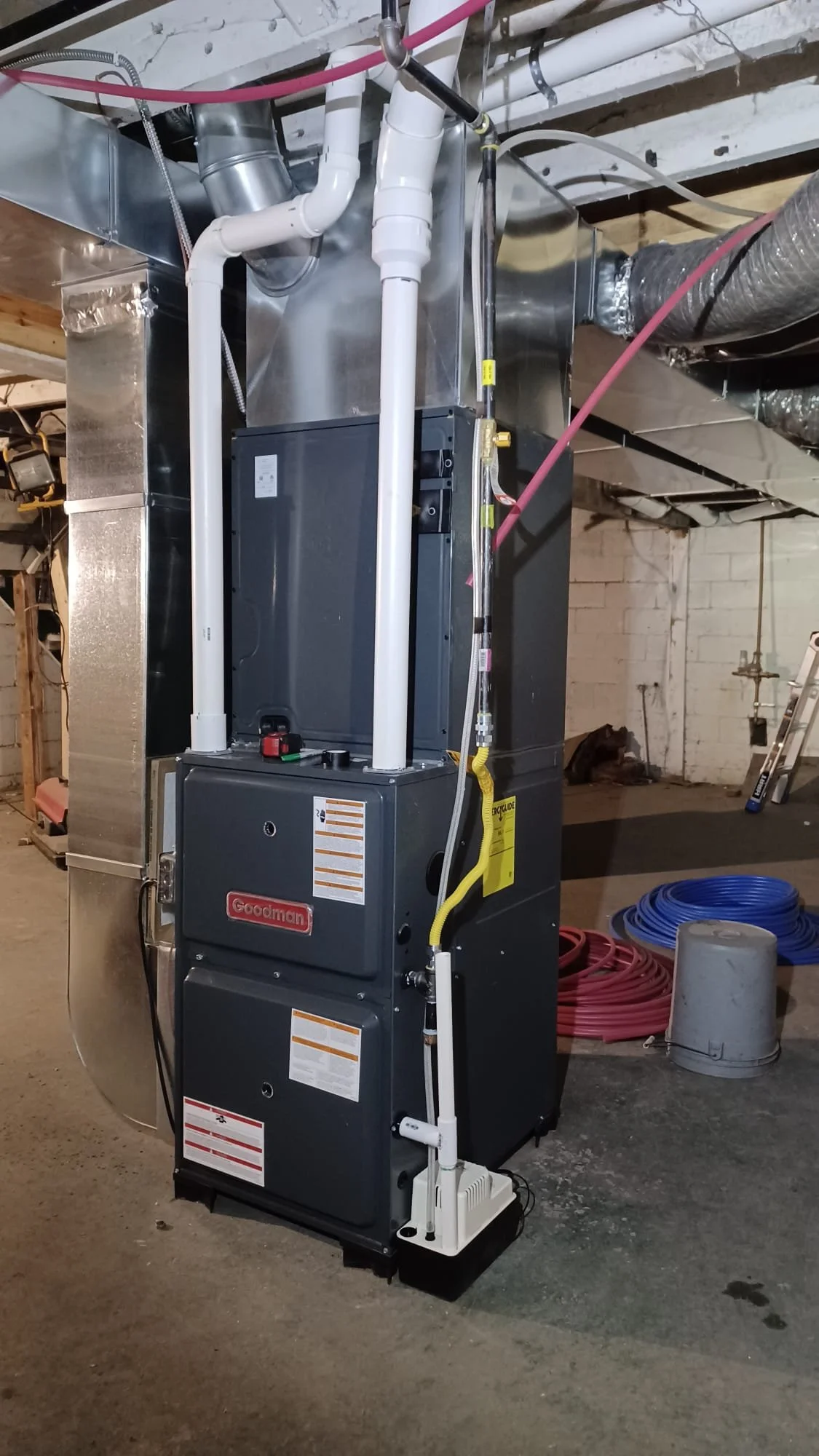 A Goodman furnace with ductwork, pipes, and electrical wires installed in a basement or utility room.