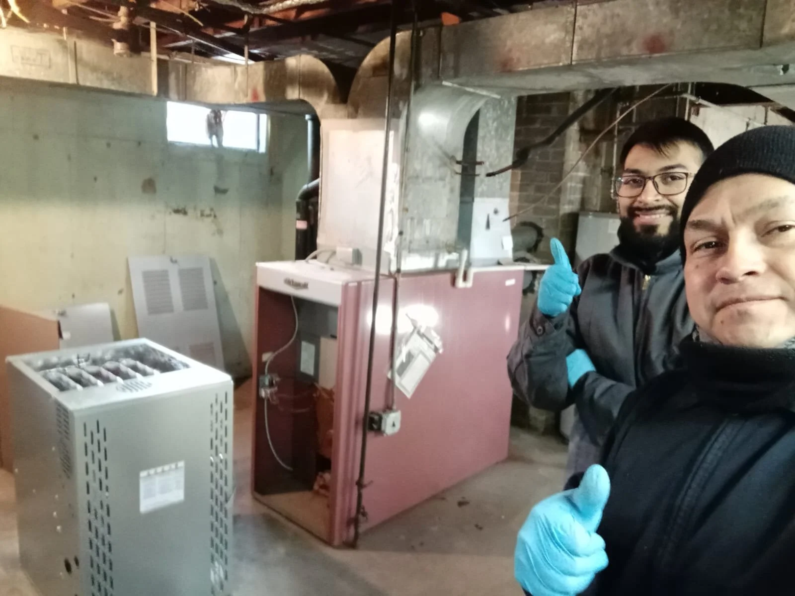 Two men inside an industrial or basement setting, standing next to large machinery, both wearing jackets and blue disposable gloves, smiling at the camera, with one giving a thumbs-up.