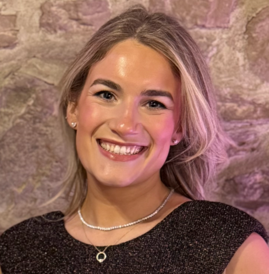 A smiling woman with shoulder-length blonde hair, wearing earrings, a pearl necklace, a layered gold necklace with a circular pendant, and a sparkly black top, standing against a stone wall background.