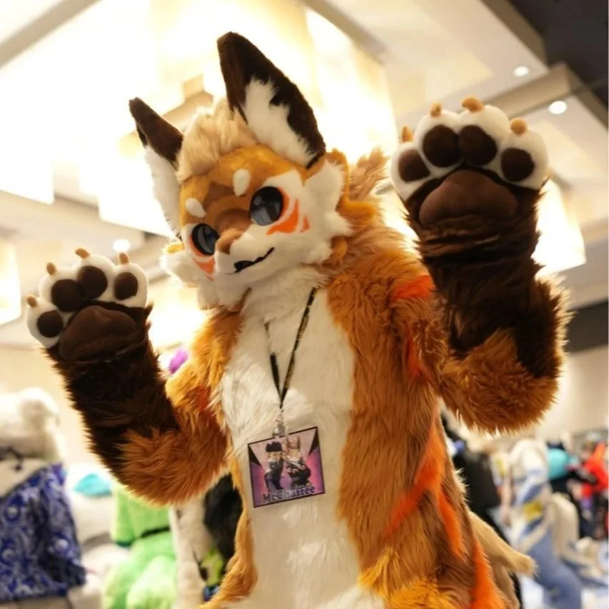 Person in a fursuit resembling a fox or coyote, with large eyes, a smiling face, and big paws with paw pads, standing indoors.