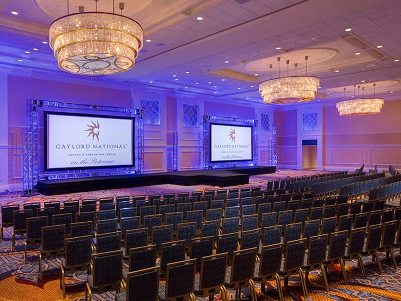 Empty conference room with two large screens displaying the Gaylord National logo, surrounded by rows of chairs and chandeliers overhead.