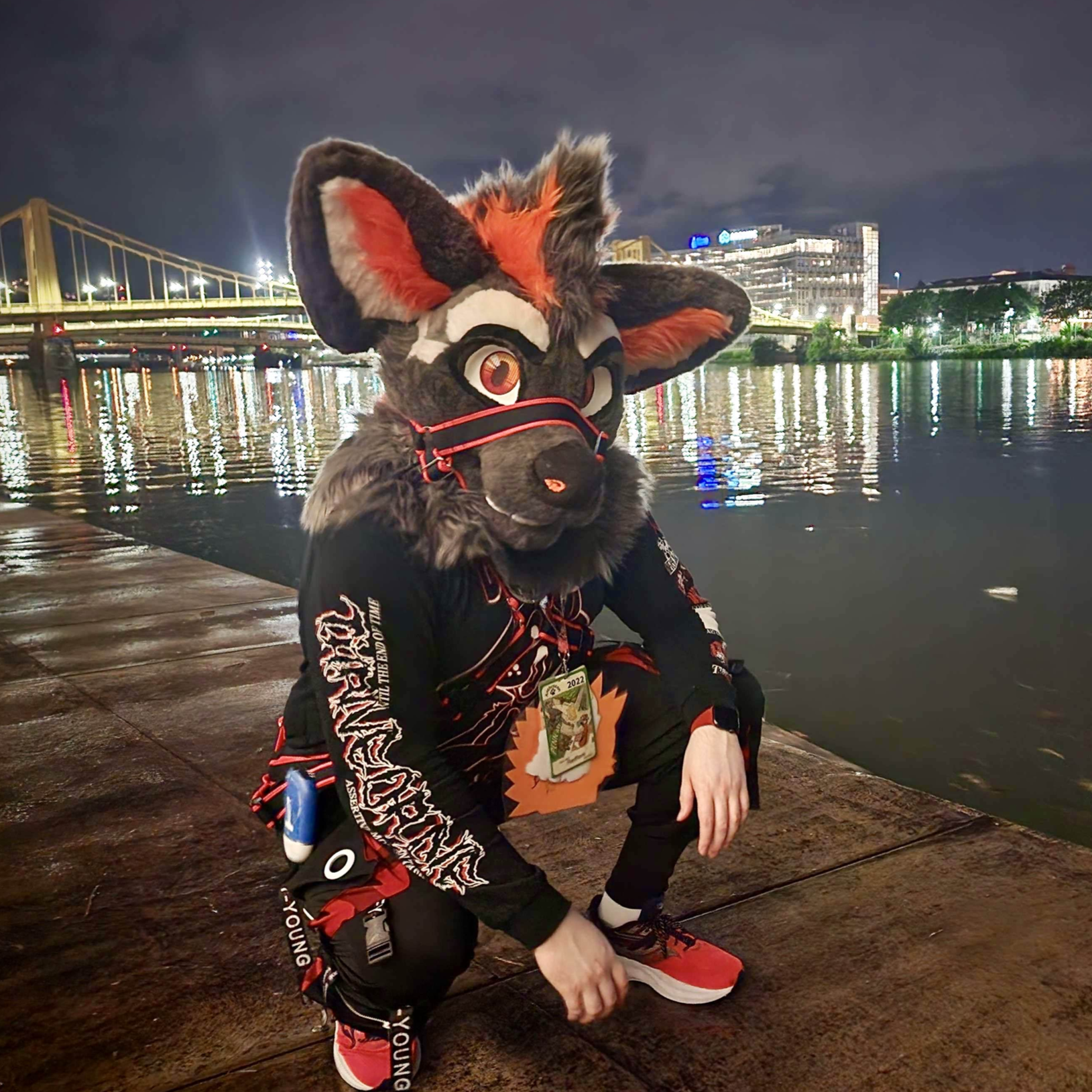 Person in a colorful animal costume crouching on wet pavement near a river at night, with city lights and a bridge reflected in the water.