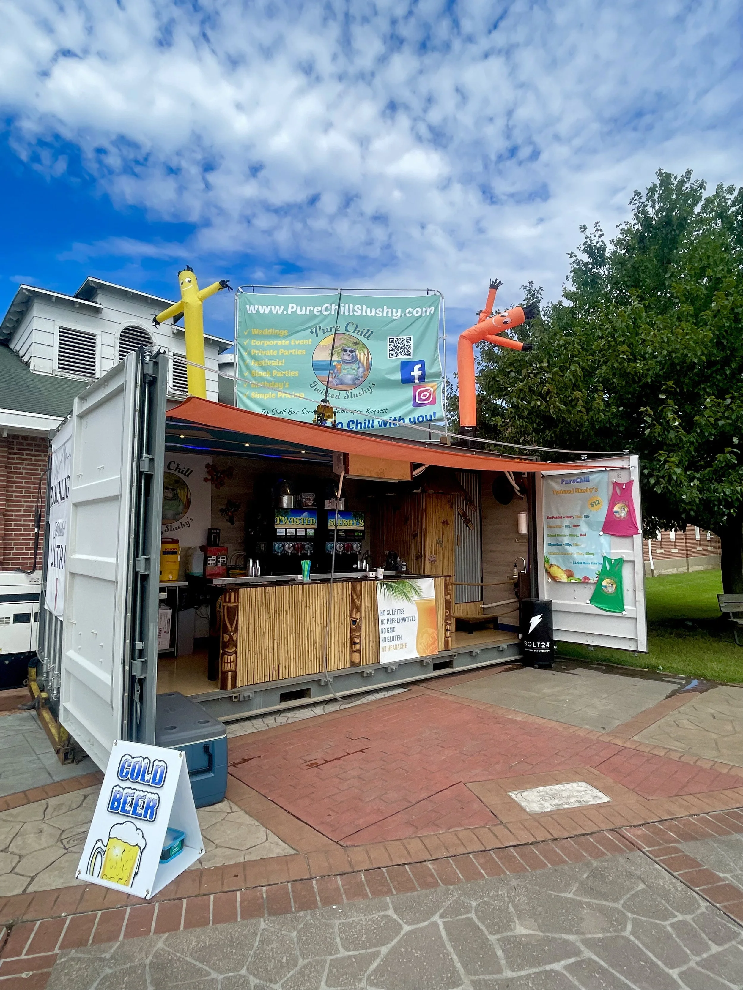 A small mobile stand with a menu sign advertising cold beer, located outdoors on a sidewalk in front of a building with a sign for Pure Chill Slushy. The stand has two large inflatable tube men, one yellow and one orange, dancing above it. There is a tree on the right side of the image and a partly cloudy sky in the background.