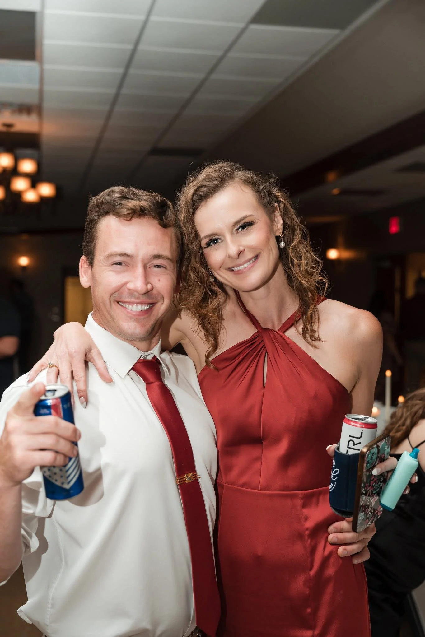A man and a woman smiling at a party, holding drinks and a phone, dressed in formal attire with dark hair and curly hair, in a dimly lit room with other people and decorations.