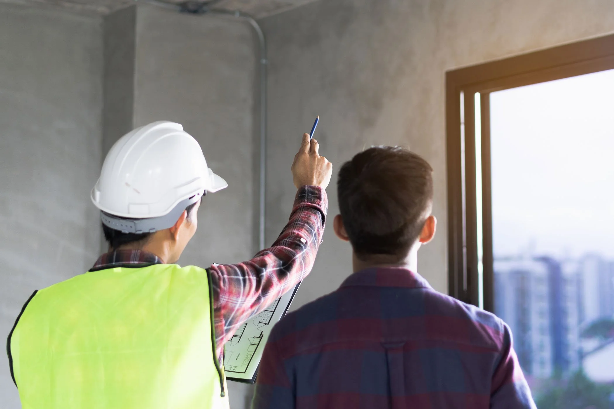 Construction worker in a yellow safety vest and white hard hat shows blueprints to a man at a construction site near a window with a city view.