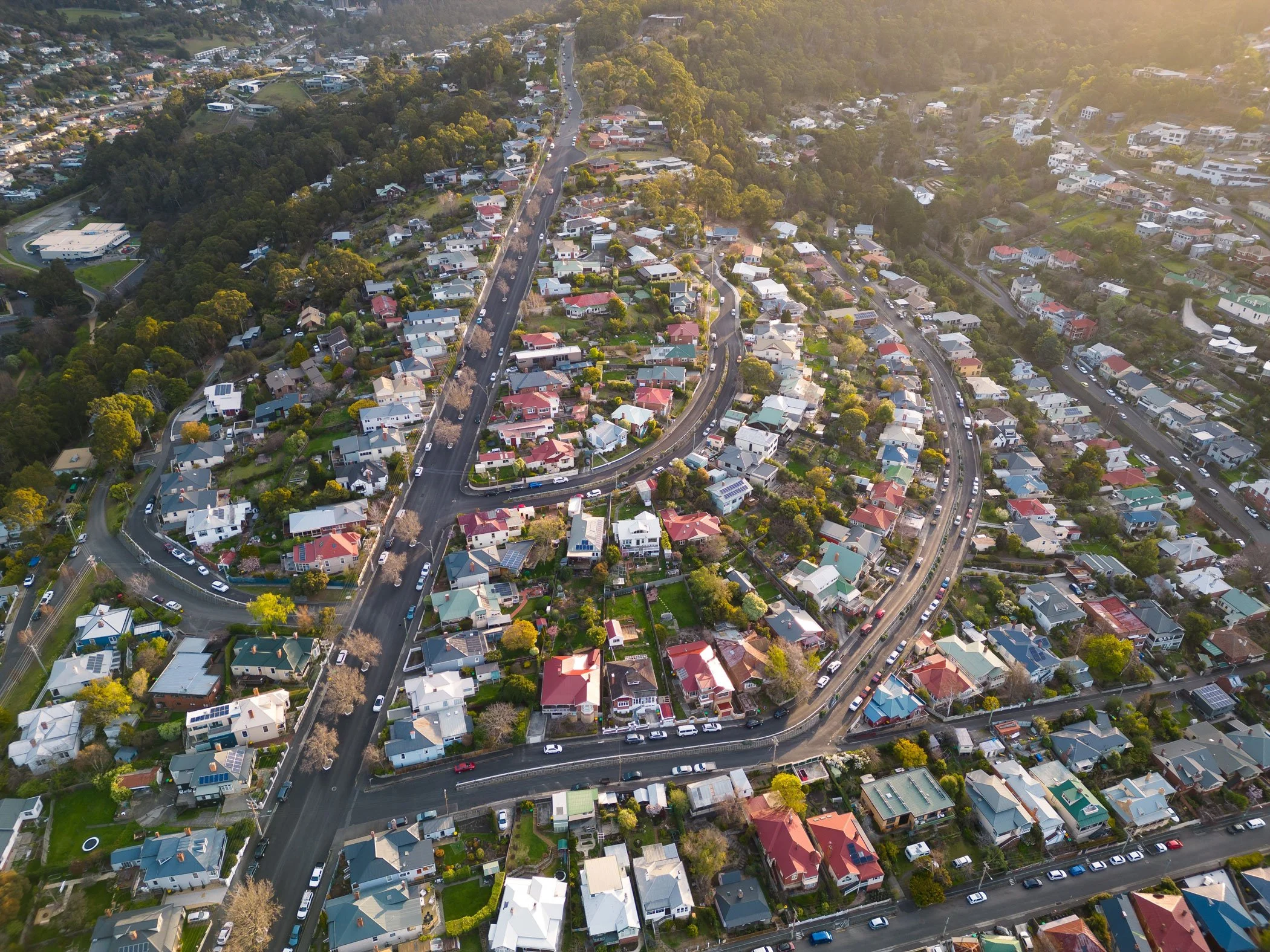 Aerial view of a residential neighborhood with houses, trees, and roads, taken during sunset with sunlight casting a warm glow.