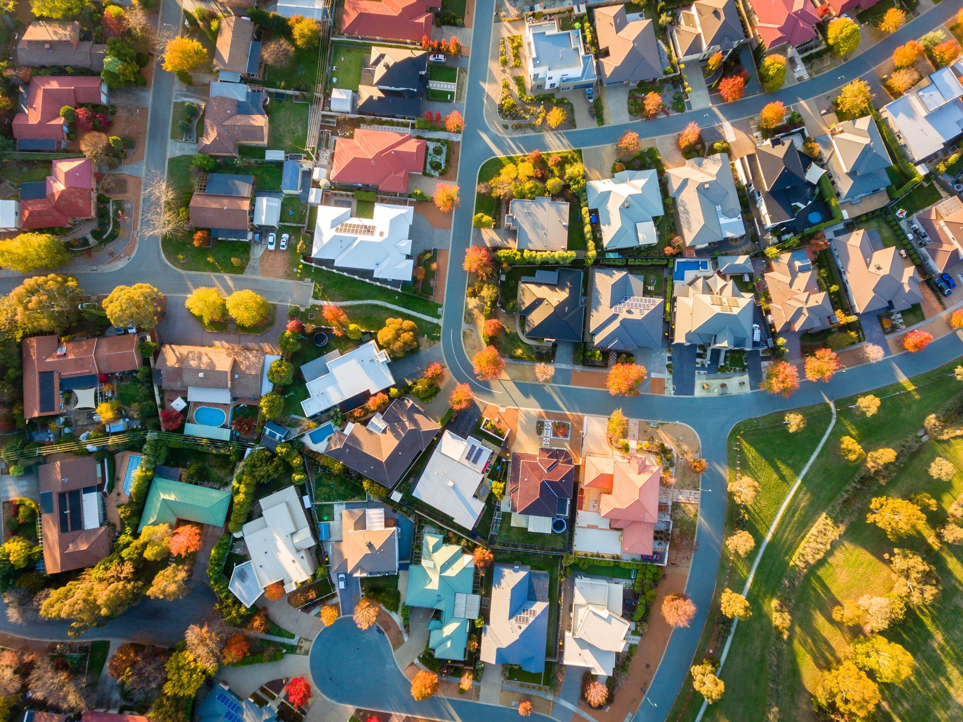 An aerial view of a suburban neighborhood during autumn, showing numerous houses with varied roof styles and colors, lined by trees with fall foliage, and intersected by winding roads.