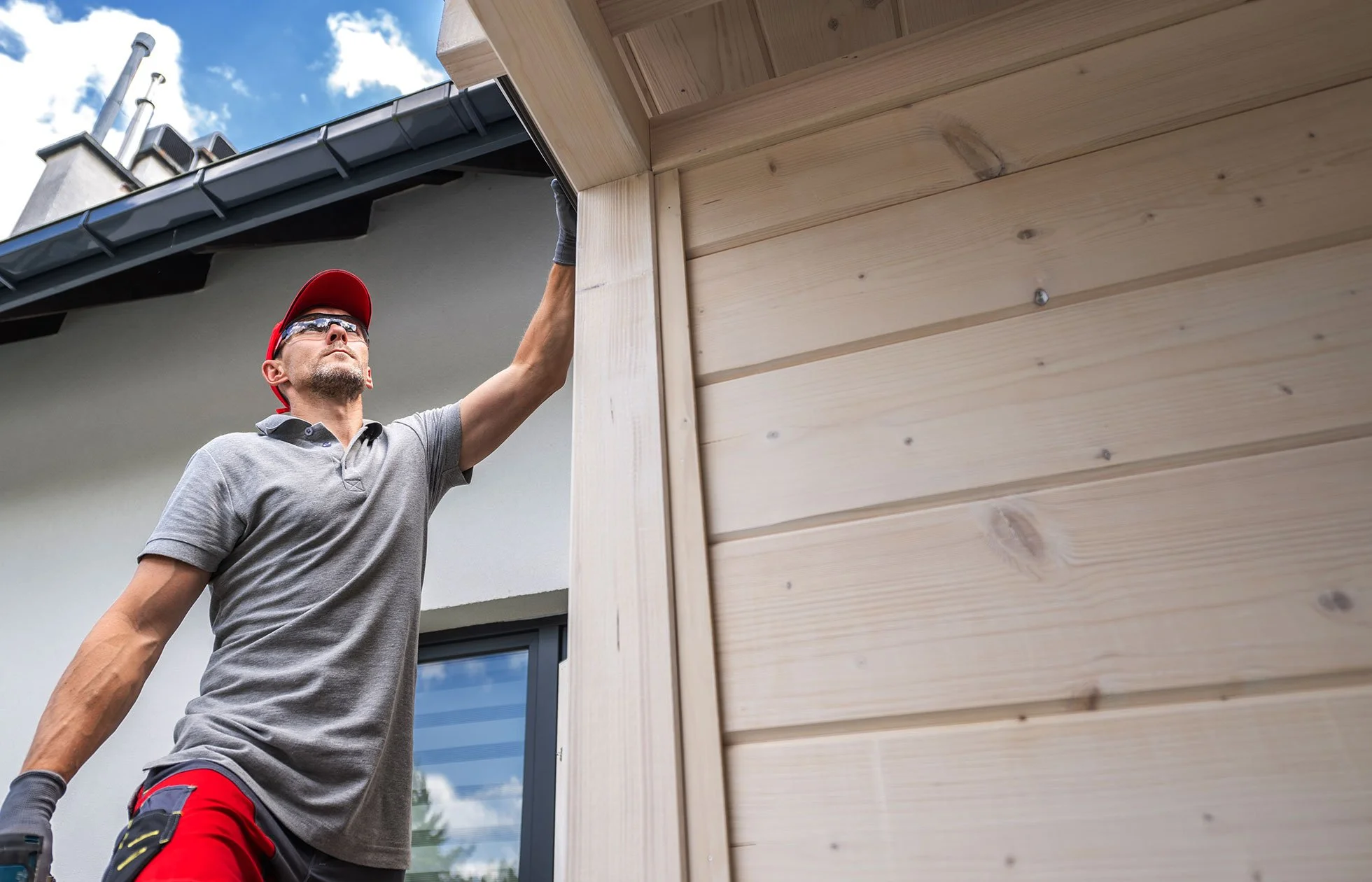 A man wearing a gray polo shirt, red cap, and sunglasses examines a wooden side of a building outdoors on a sunny day.