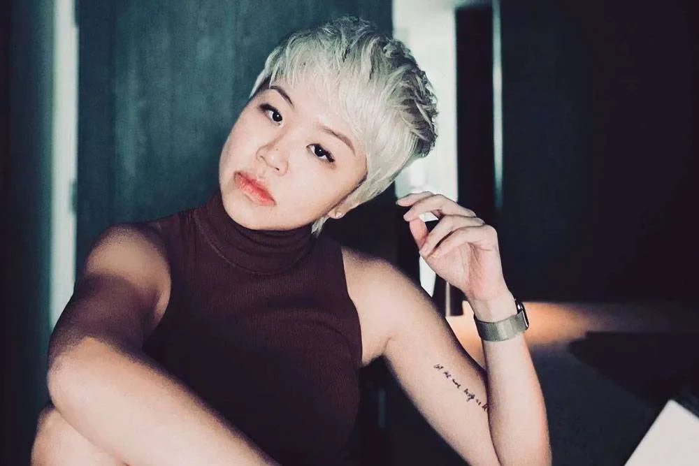 A young woman with short, platinum blonde hair wearing a sleeveless dark top, looking at the camera while adjusting her earring in a dimly lit room.