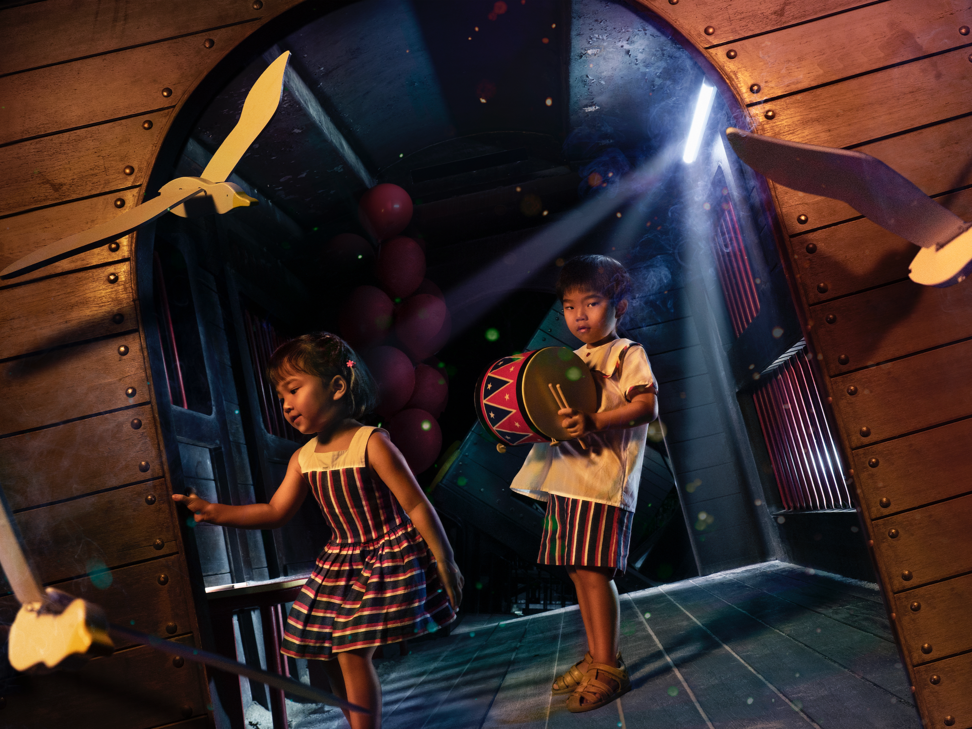 Two children, a girl and a boy, are playing in a wooden tunnel with adjustable shields and hanging balloons. The girl is near the entrance, and the boy is holding a drum and drumsticks, facing the camera. The lighting creates a dramatic atmosphere with a beam of light.