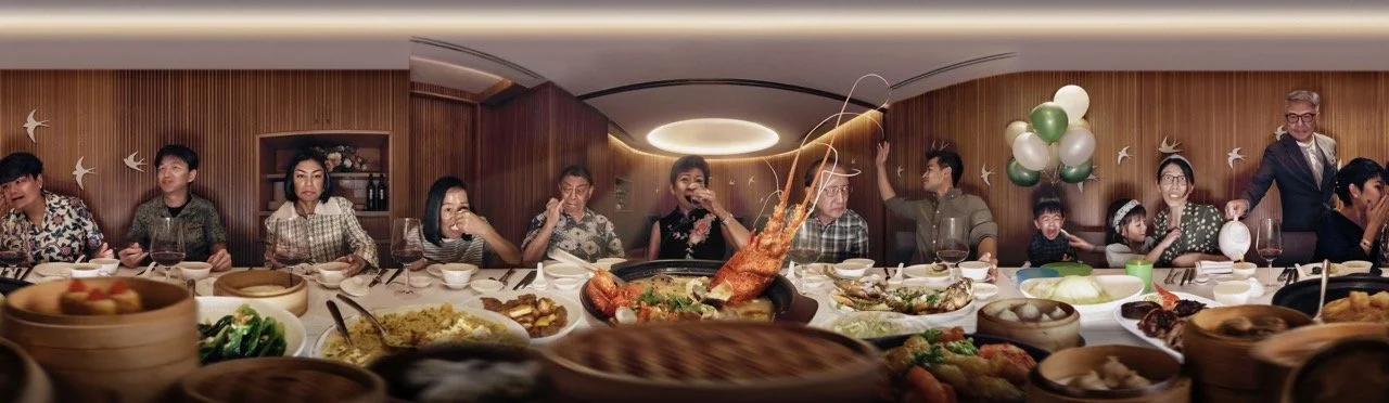 People sitting around a dinner table with a variety of dishes, a hot lobster in the center, and balloons in the background.