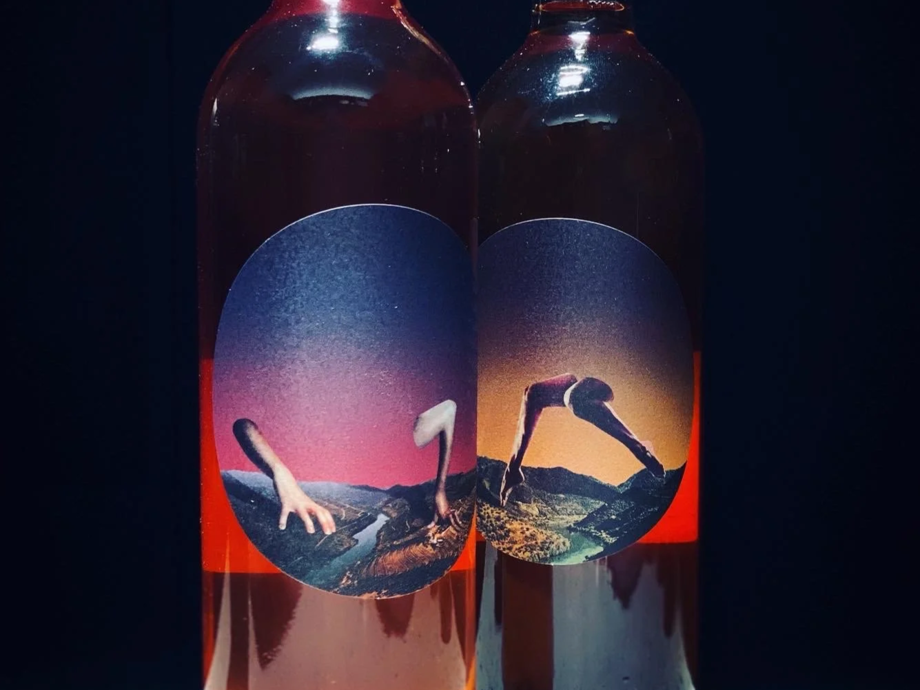 Two glass bottles with colorful labels featuring surreal landscape images and human hands, sitting on a speckled white surface with a dark background.