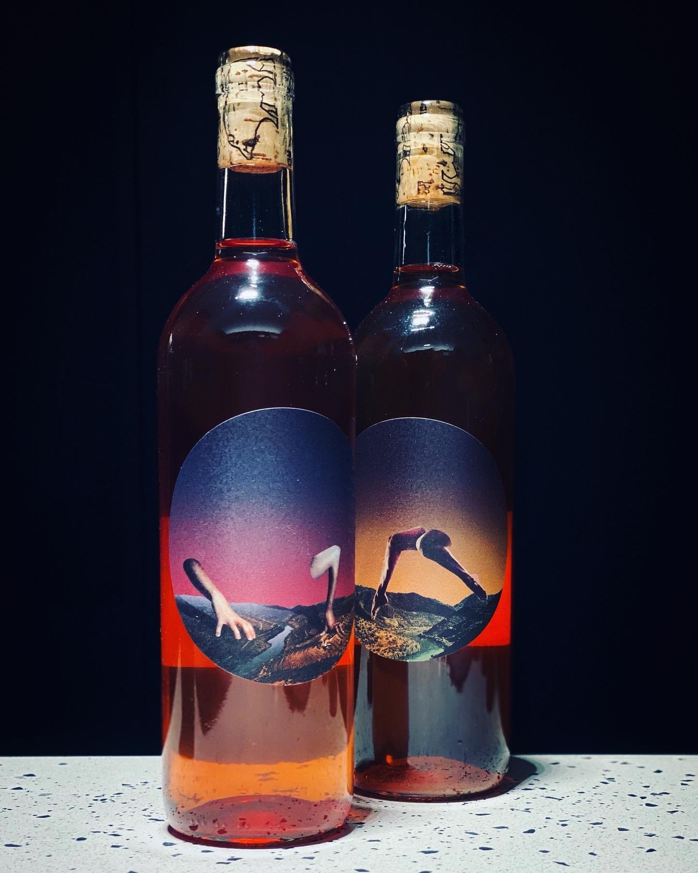 Two glass bottles with colorful labels featuring surreal landscape images and human hands, sitting on a speckled white surface with a dark background.