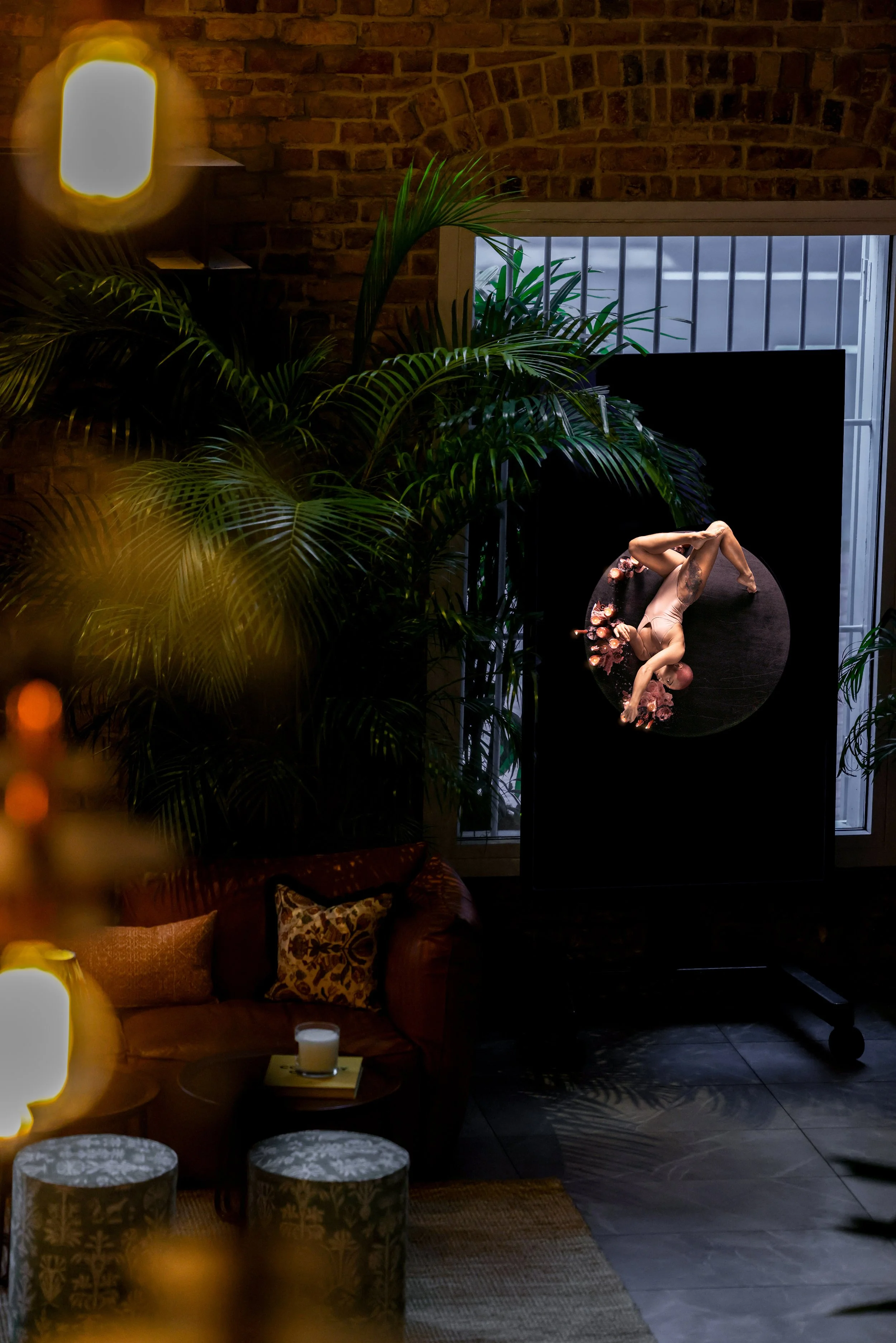 A woman stretching on a round mirror or art piece, surrounded by large green plants and low lighting in a cozy ambient room.