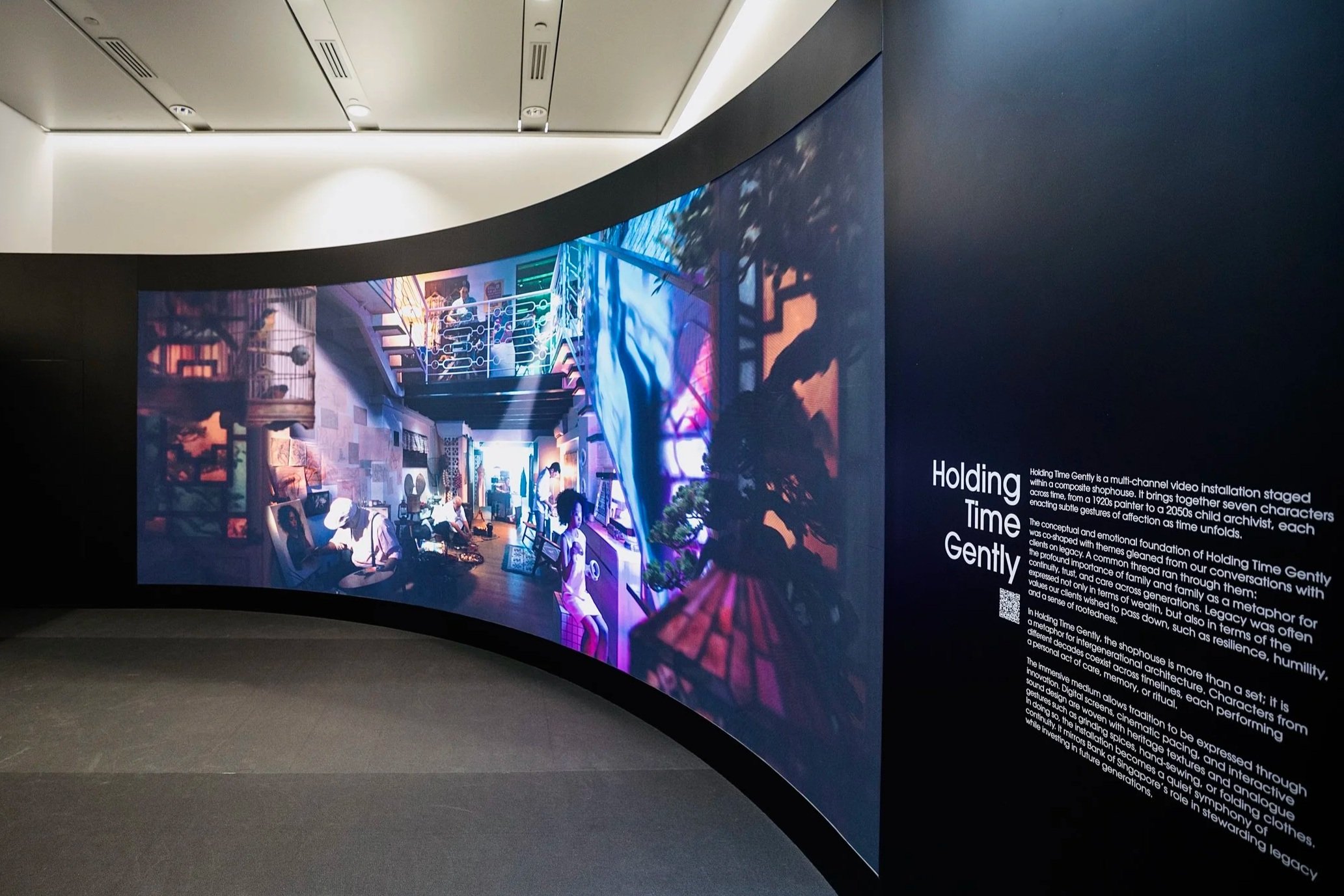 A curved digital display in a gallery or museum shows a colorful digital scene with people and artwork. Adjacent to the screen, a black wall has white text explaining the exhibit.