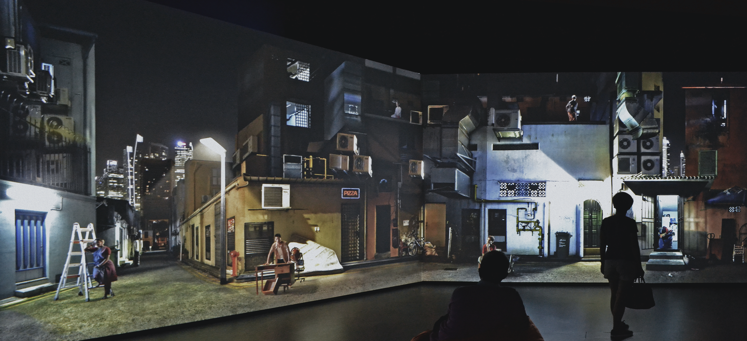 Nighttime city scene with multiple buildings, air conditioning units, and a few people walking; one person sitting and another standing near a corner, with a person in the background on a balcony.