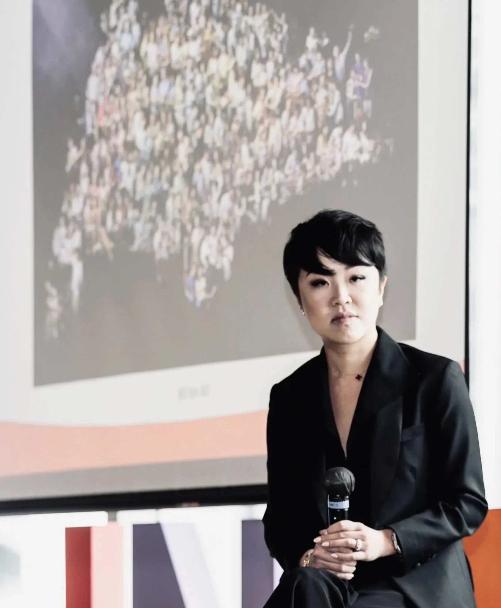 A woman with short black hair, dressed in black, sitting with a microphone in her hand, in front of a large screen displaying a crowd photo.