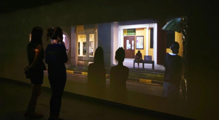 People watching a projection of a street scene with benches and storefronts on a wall in a dark room.