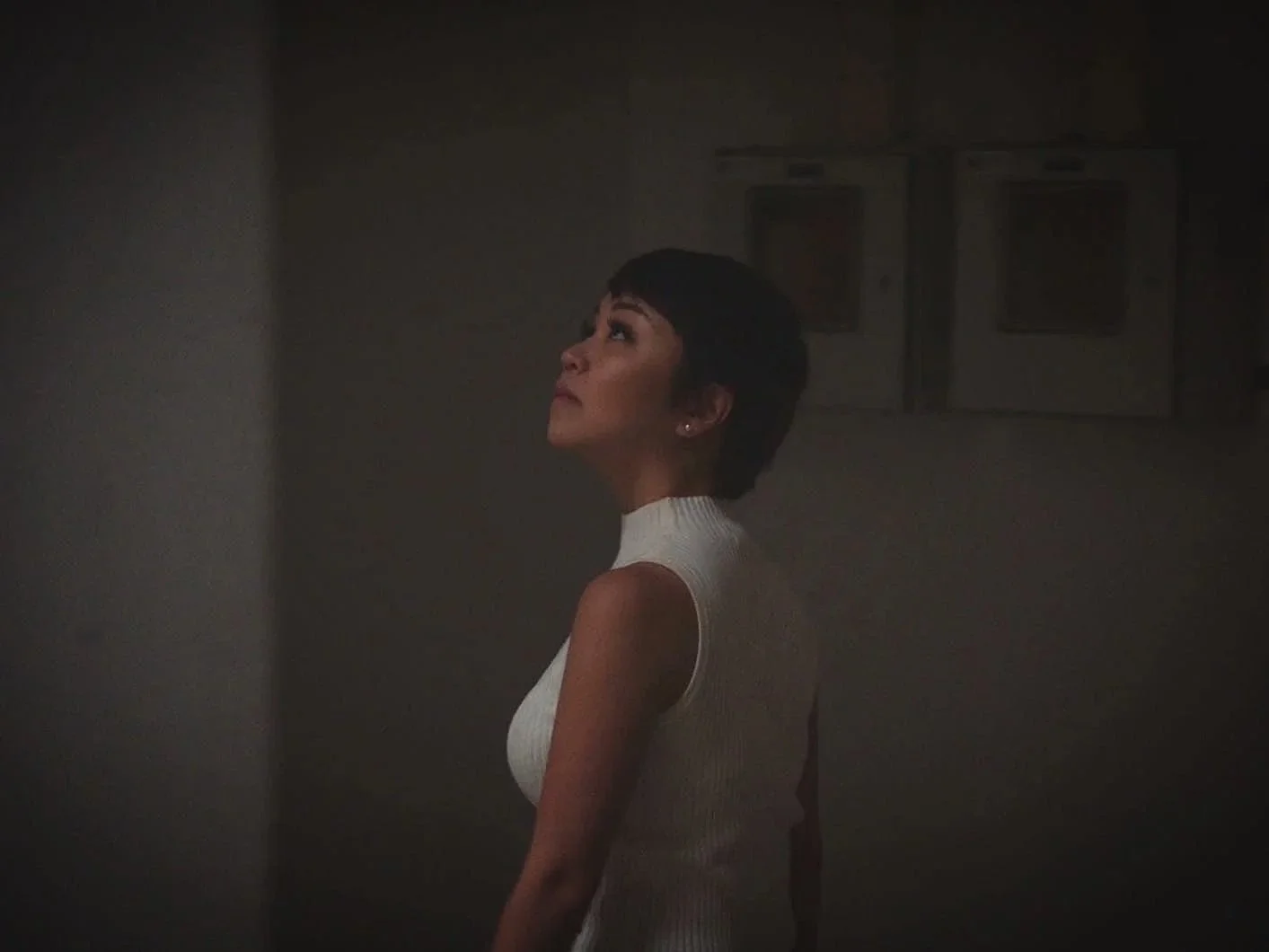 A woman with short dark hair, wearing a white sleeveless turtleneck, looking upward in a dimly lit room with two laundry appliances in the background.