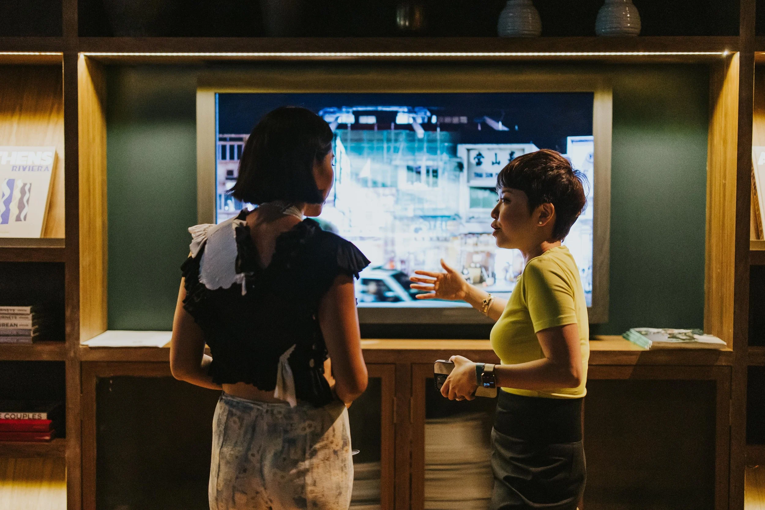 Two women having a conversation in front of a television in a cozy room with bookshelves.