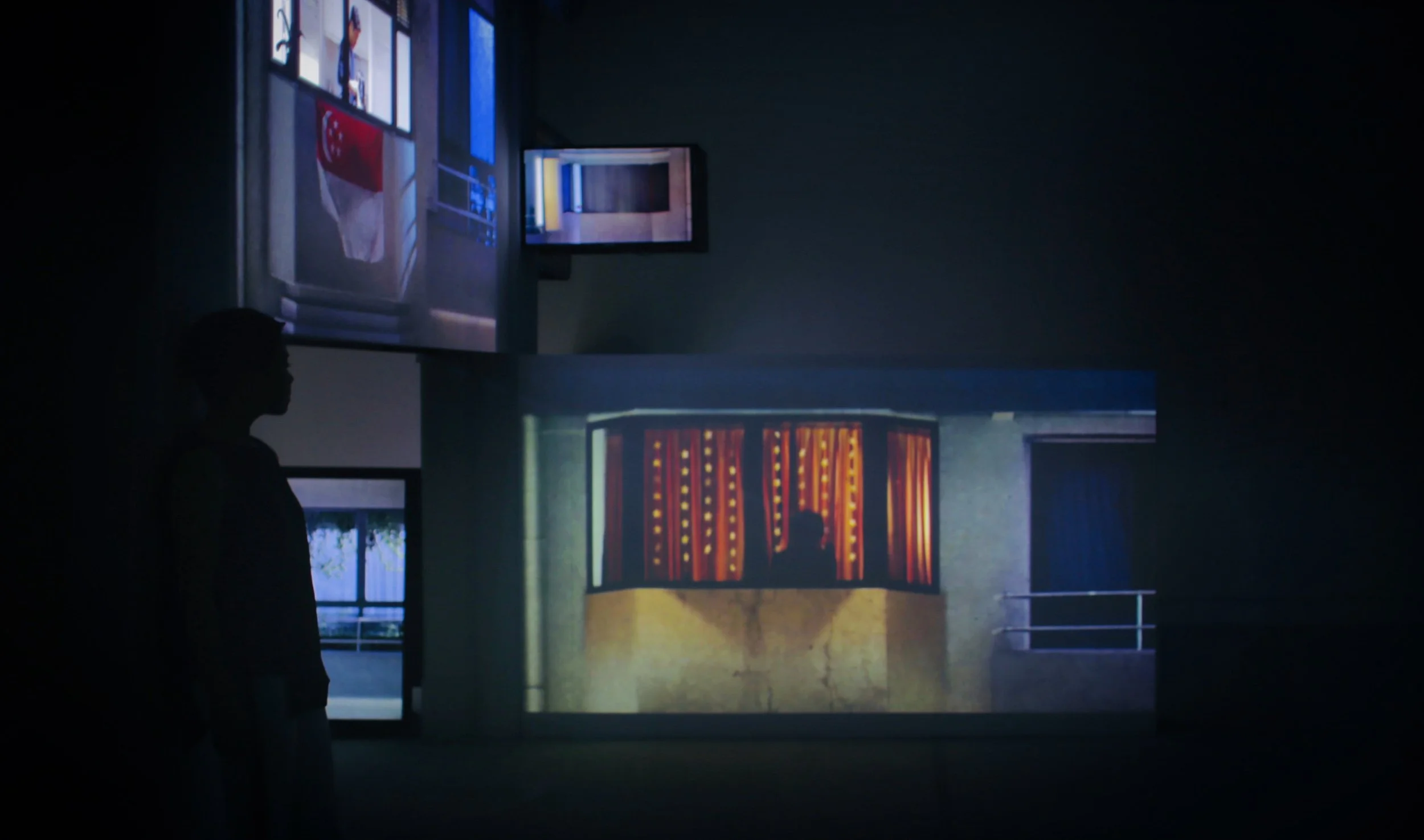 Silhouette of a person standing in a dark room with multiple screens displaying various images, including a person behind a stage with curtains.