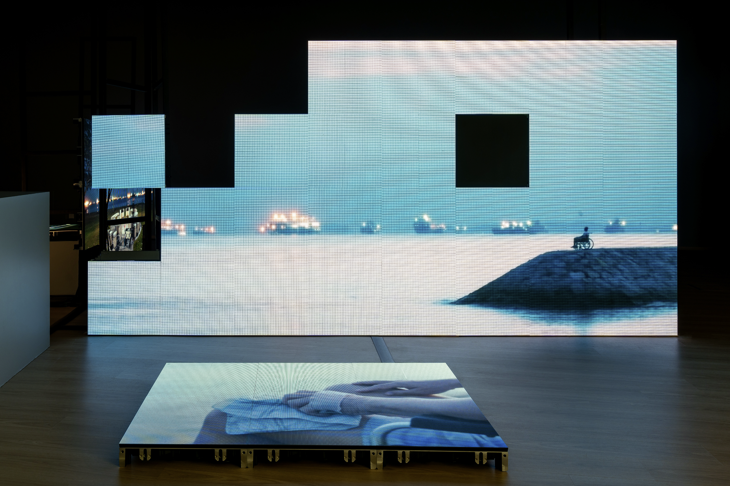 A pixelated digital display art installation showing a person in a wheelchair on a rocky hill by the water, with abstract city lights in the background.