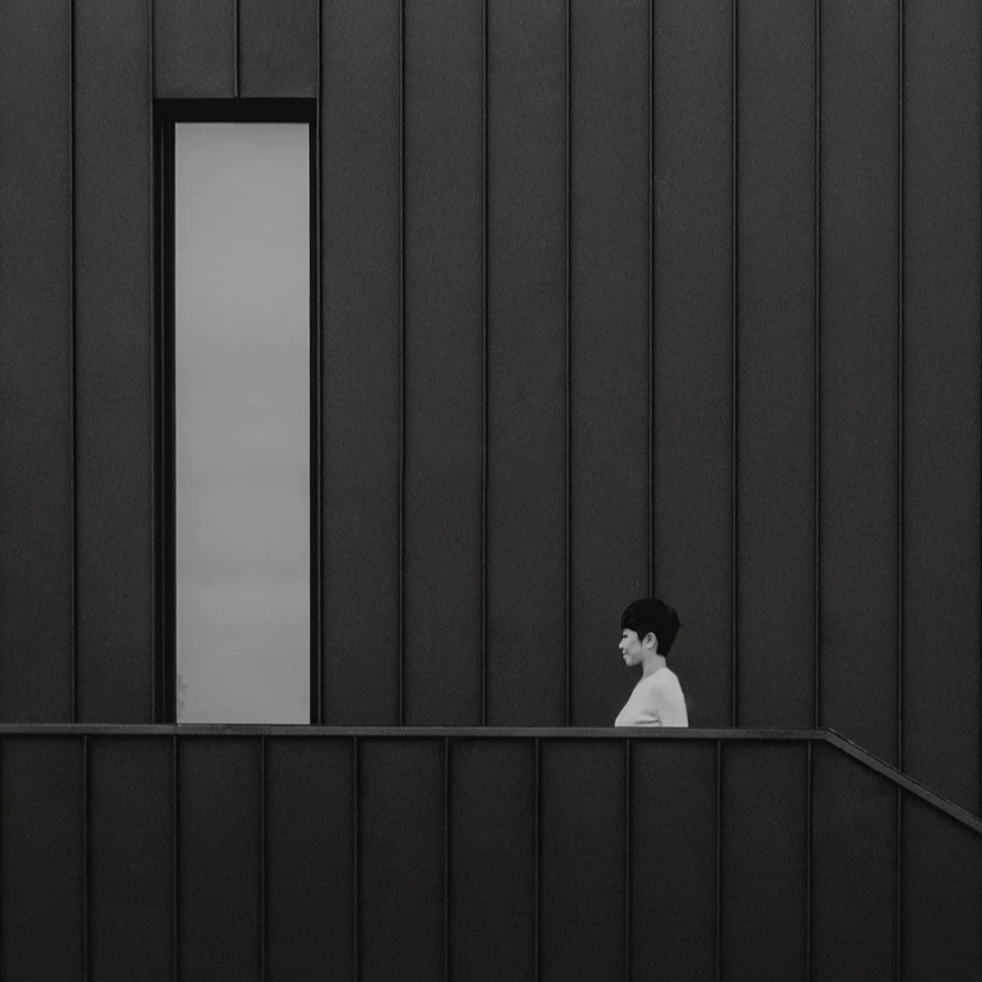 A woman with short dark hair and a white top standing on a black urban balcony against a dark building with vertical panels and a tall window.