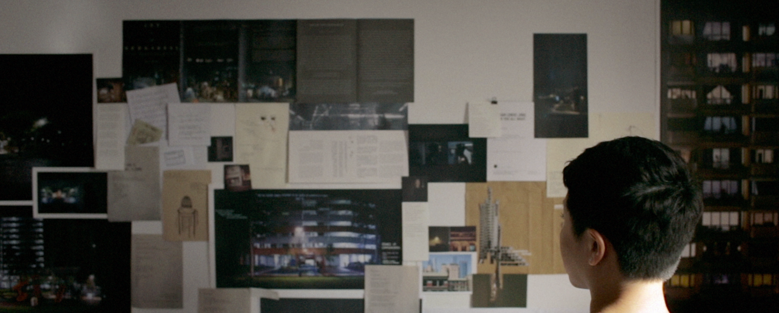 A person with short dark hair looking at a bulletin board filled with various papers, photos, and sketches, in a room with bookshelves in the background.