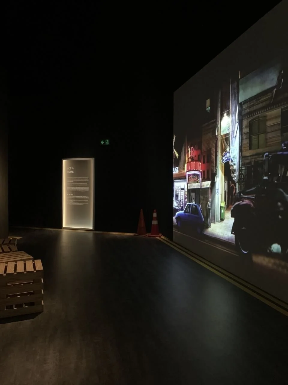 Dark interior space with a large illuminated photograph of a street scene on the right wall, featuring cars and buildings. There are two traffic cones near the corner and a lit informational display on the left side.