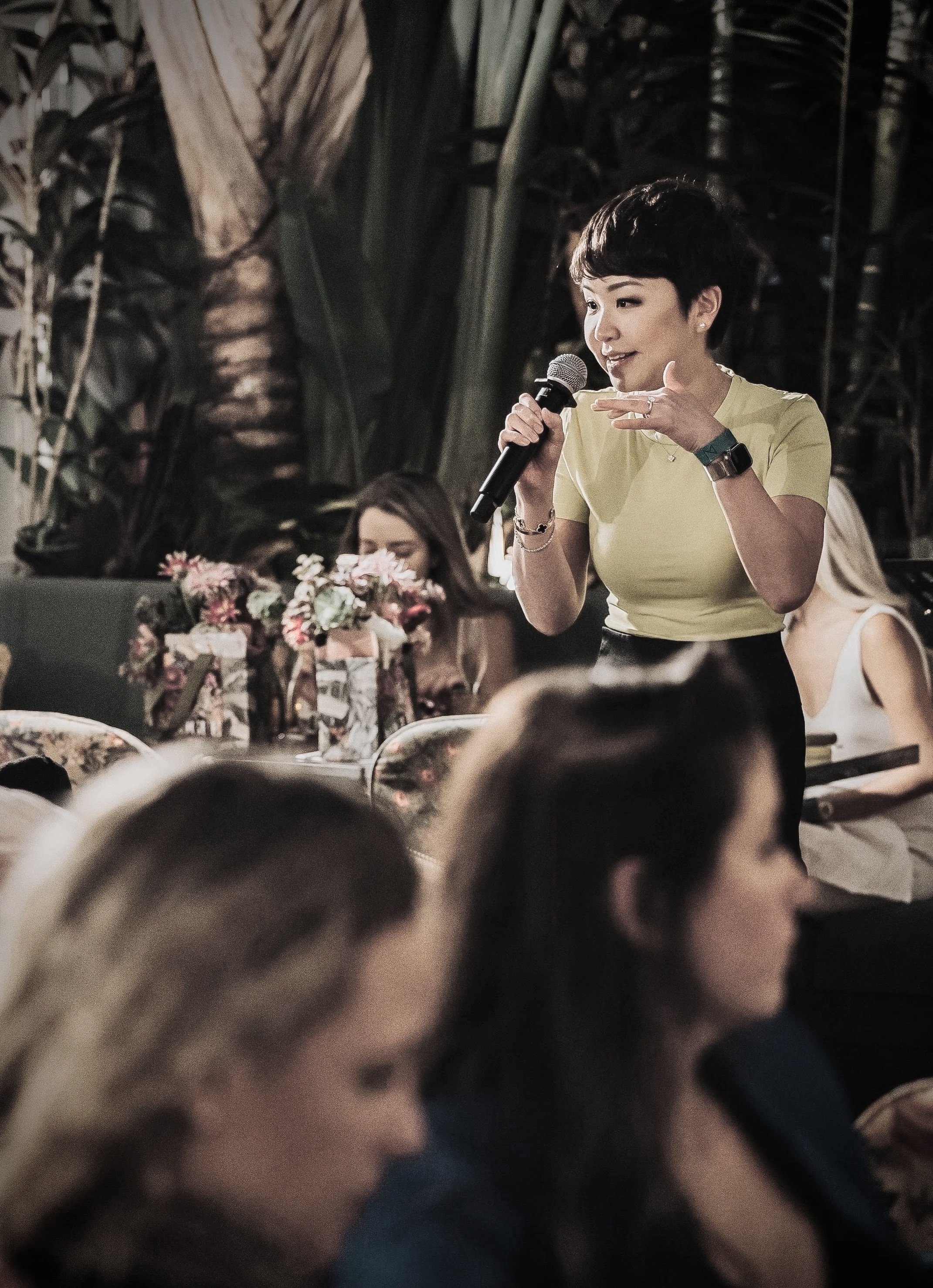 A woman in a yellow shirt speaking into a microphone during an event, with others seated at a table decorated with flowers, in a setting with tropical plants.