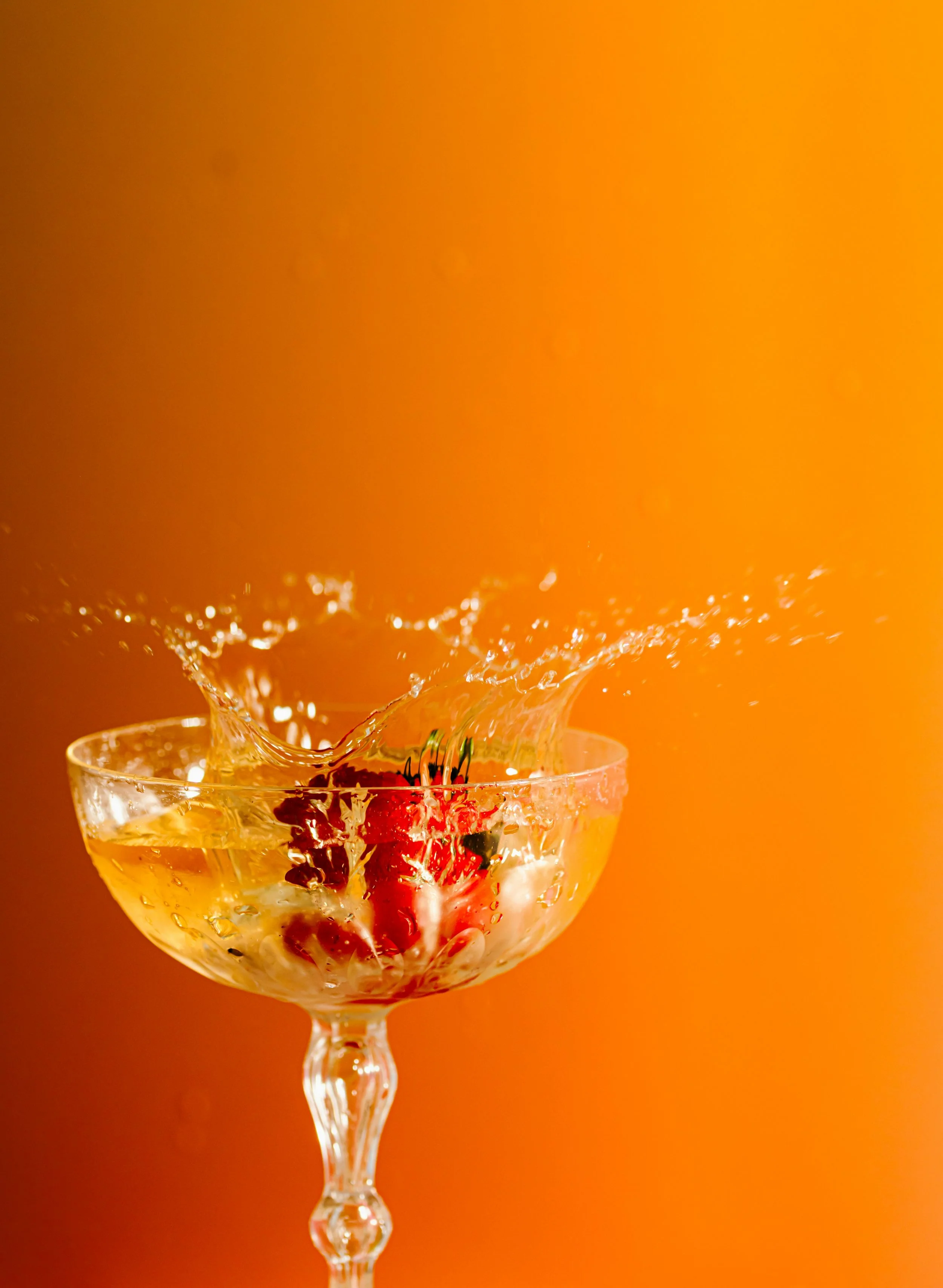 A glass coupe filled with lush Vera Pinot Gris with a splash of liquid and berries inside against an orange background.