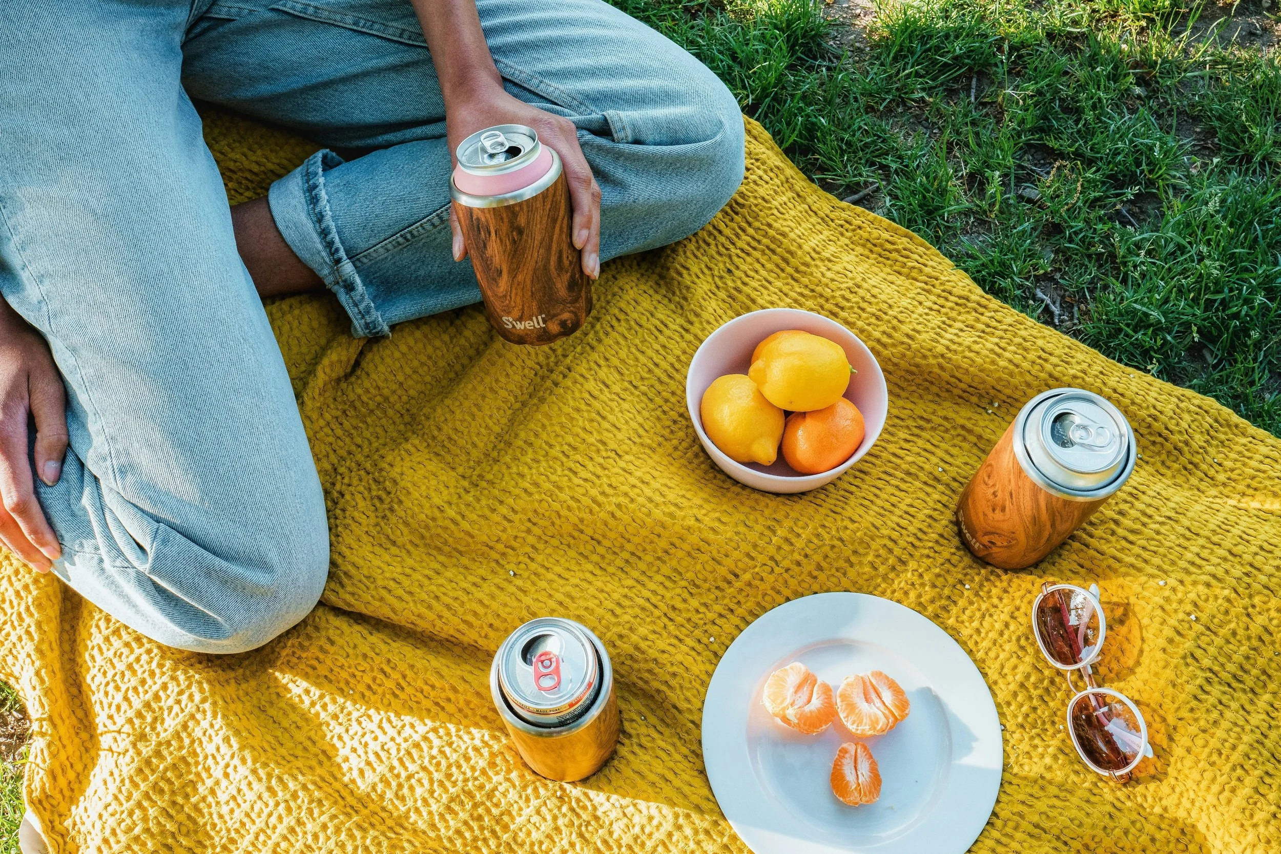Close-up of a picnic setup on a yellow blanket with a person's legs visible, holding a can of Swell drink. There are bowls of lemons and oranges, a plate with peeled orange segments, sunglasses, and additional cans of drink placed on the blanket, wit