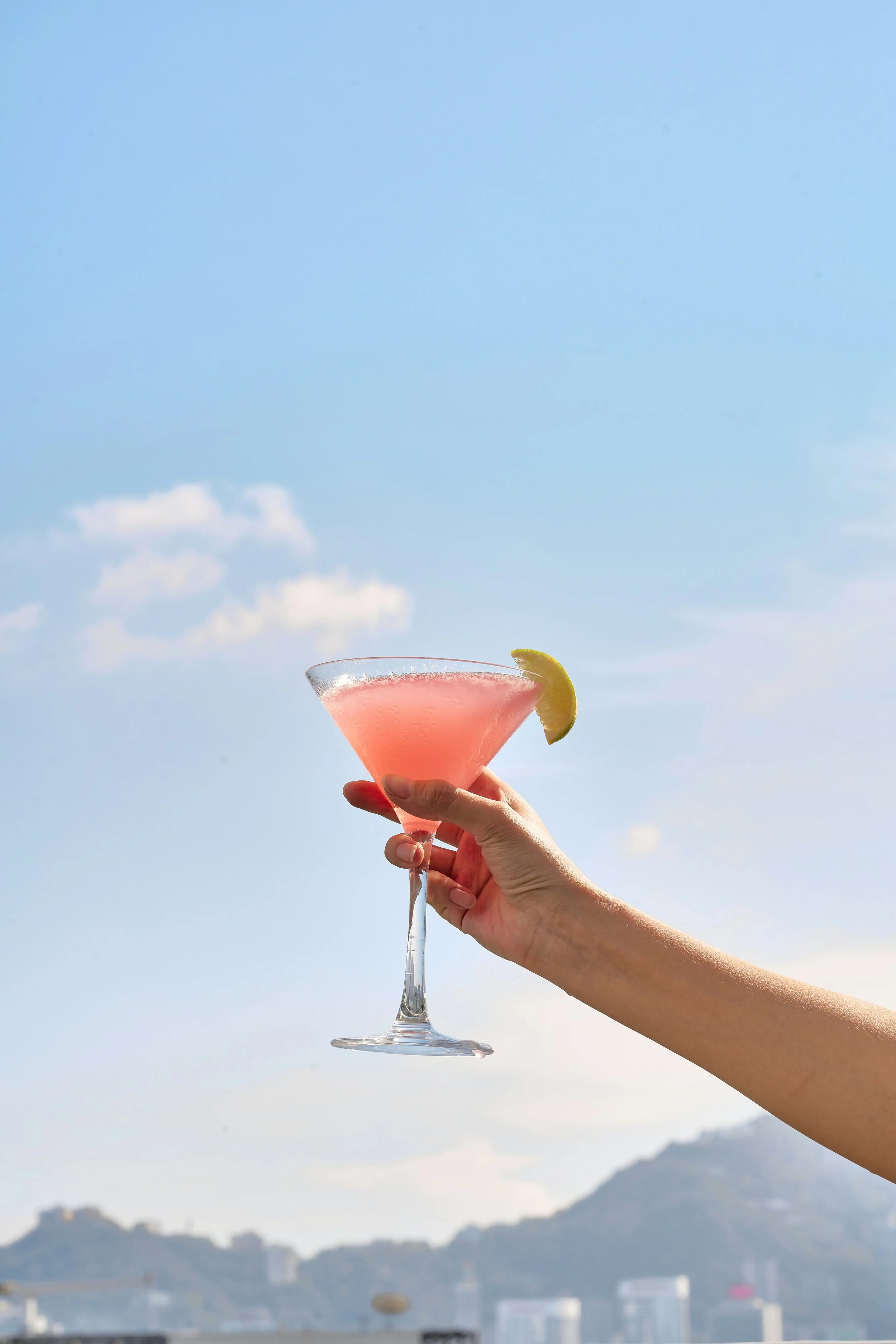 A hand holding a pink cocktail with a lemon wedge garnish against a blue sky with some clouds and a cityscape with hills in the background.