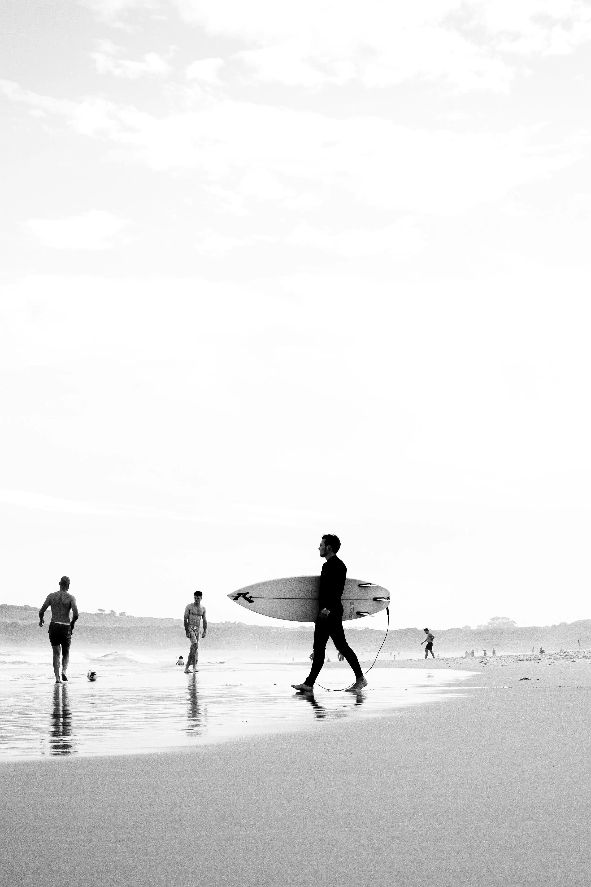 Black and white photo of a man in a suit walking along a beach with a surfboard, while other people are in the background playing with a ball and in the water.