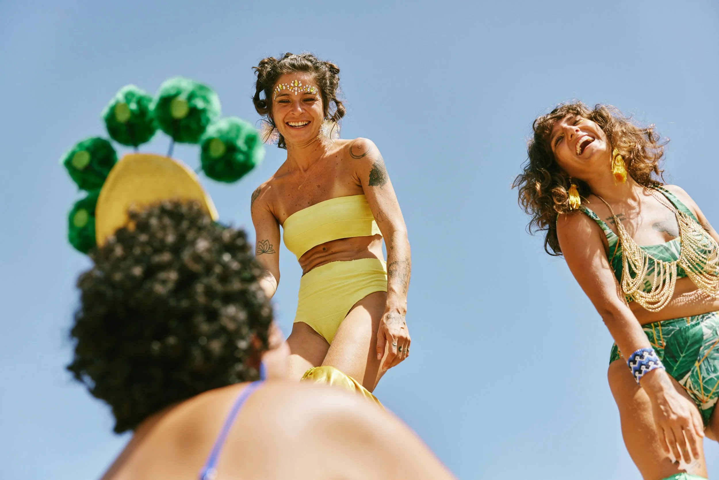 Fun in the sun with Vera Seltzers - Two smiling women dressed in bright yellow and green summer outfits, with one wearing jewelry and face paint, standing under a clear blue sky.