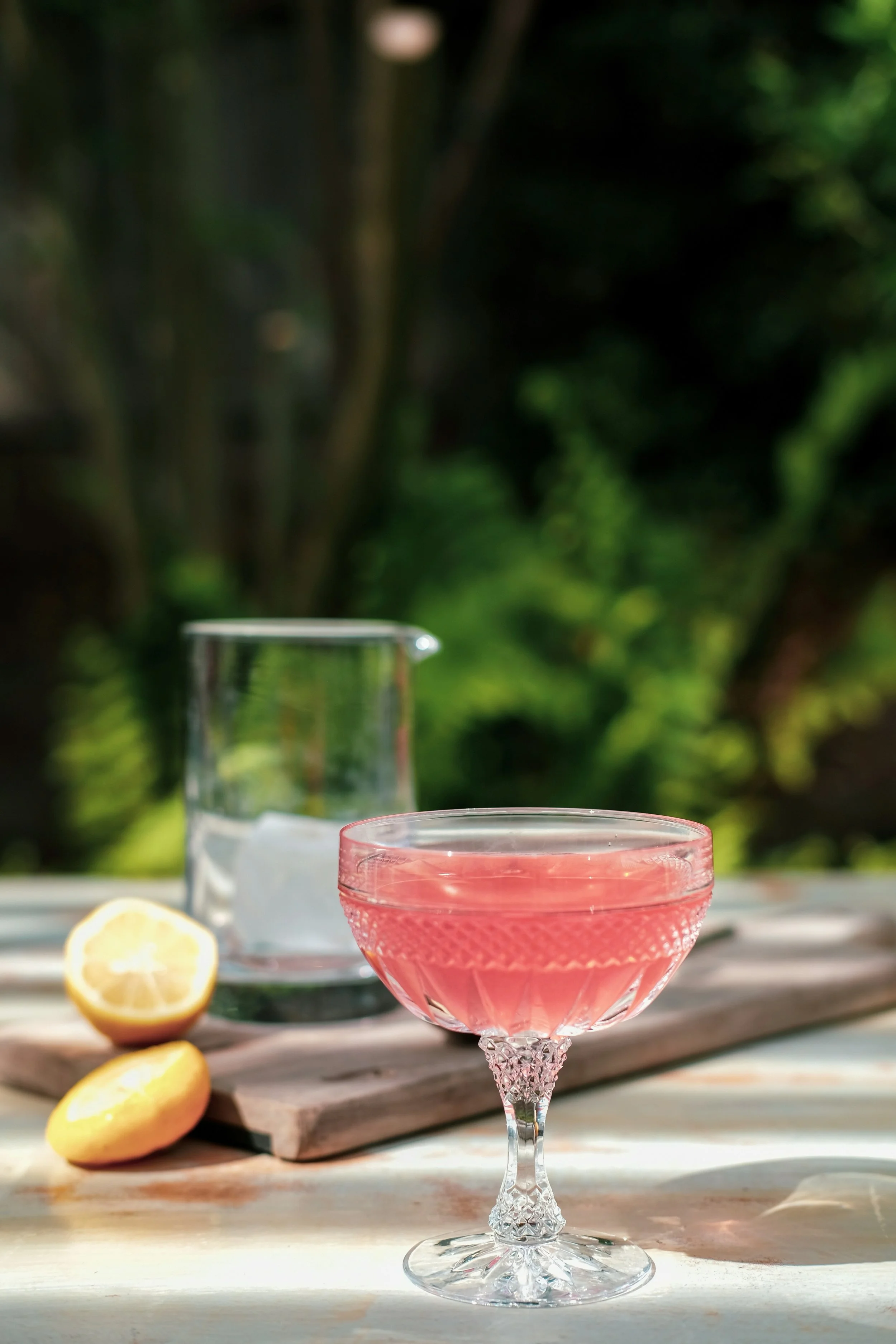 A Vera Rose cocktail in a vintage-style glass, a lemon halves, and a glass of water with ice on a wooden surface outdoors with green blurred trees in the background.