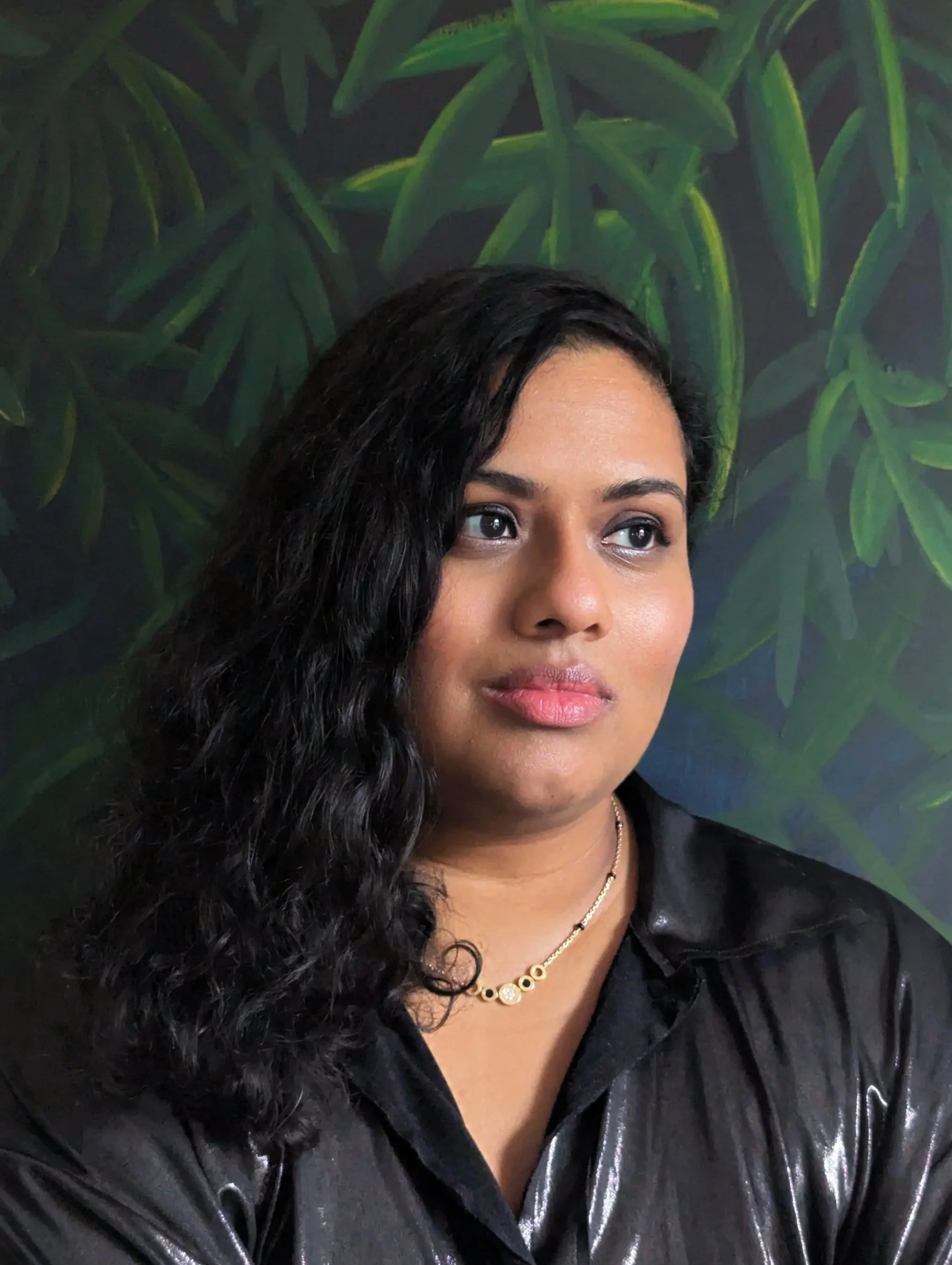 A woman with dark, curly hair and medium skin tone looking to the side. She is wearing a black leather jacket and a gold necklace, with a green leafy background.