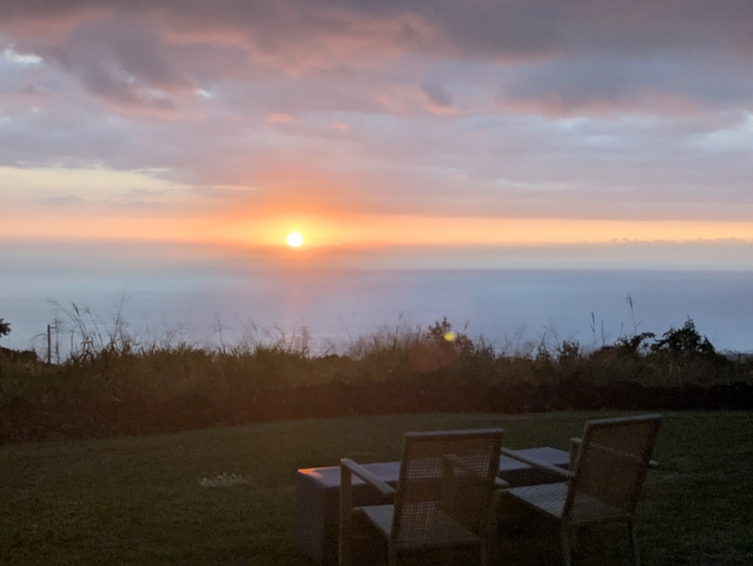 A sunset over the ocean with a partially cloudy sky, seen from a grassy area with outdoor chairs and a table.