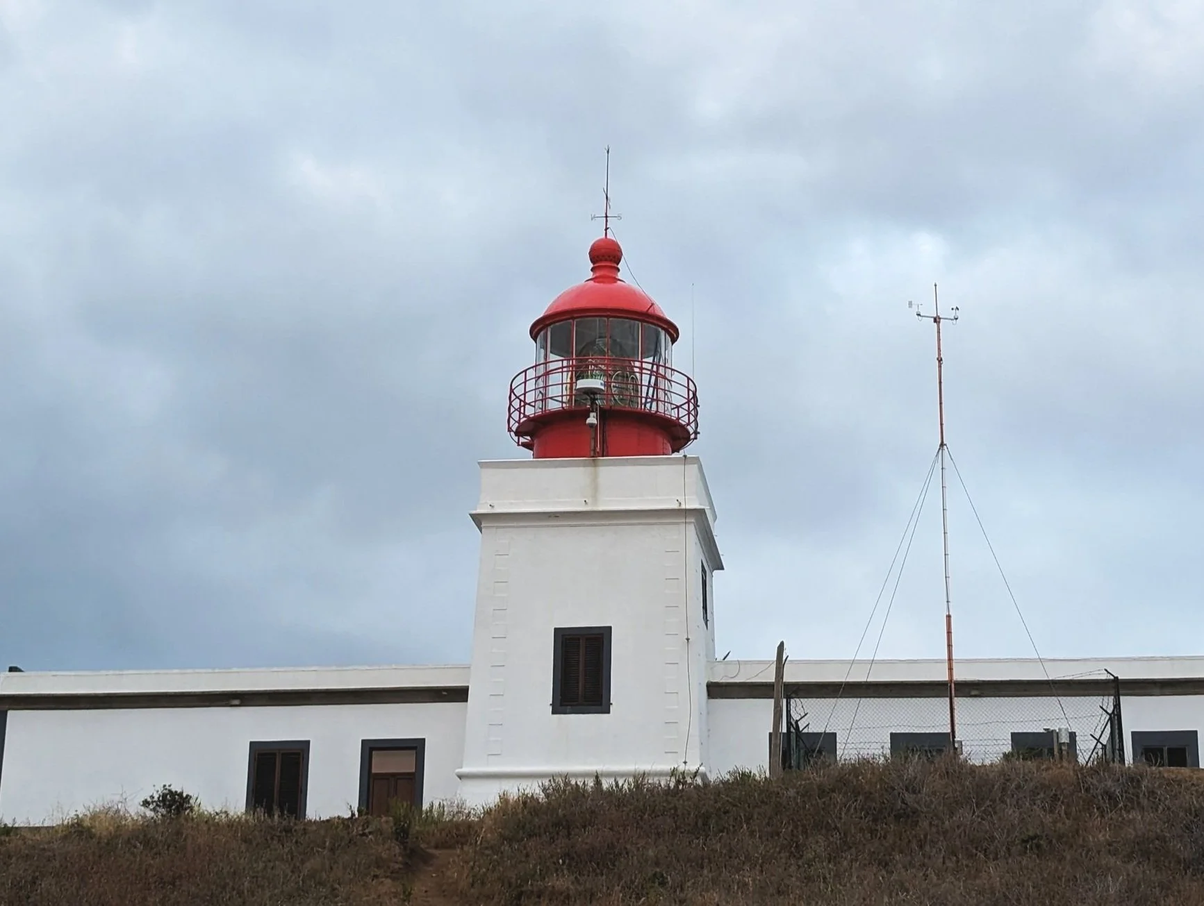 A white lighthouse with a red top and railing, situated on a grassy hillside under a cloudy sky.