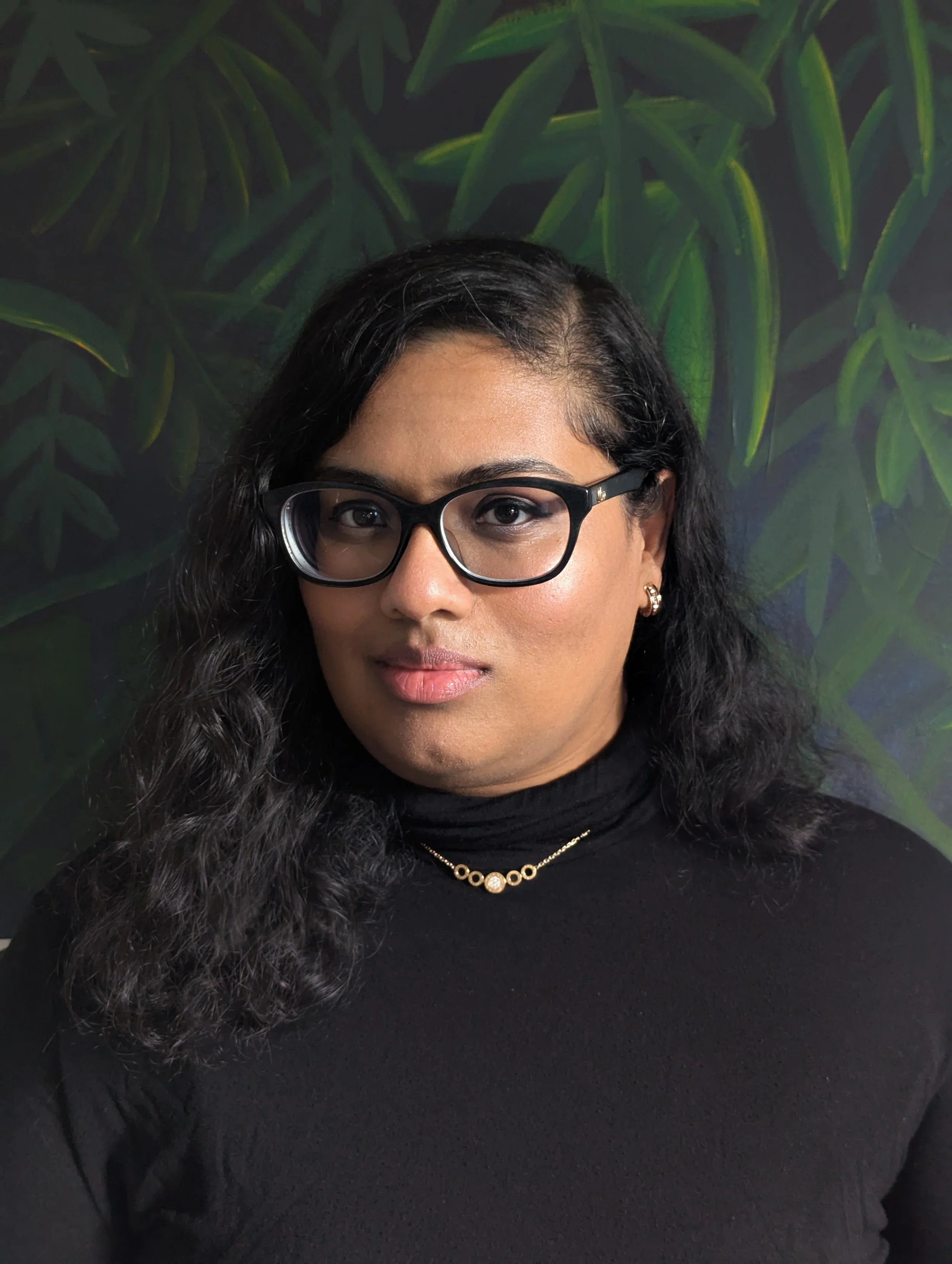 Portrait of a woman with black, curly hair, wearing glasses, a black turtleneck, and gold jewelry, standing in front of a plant-themed background.