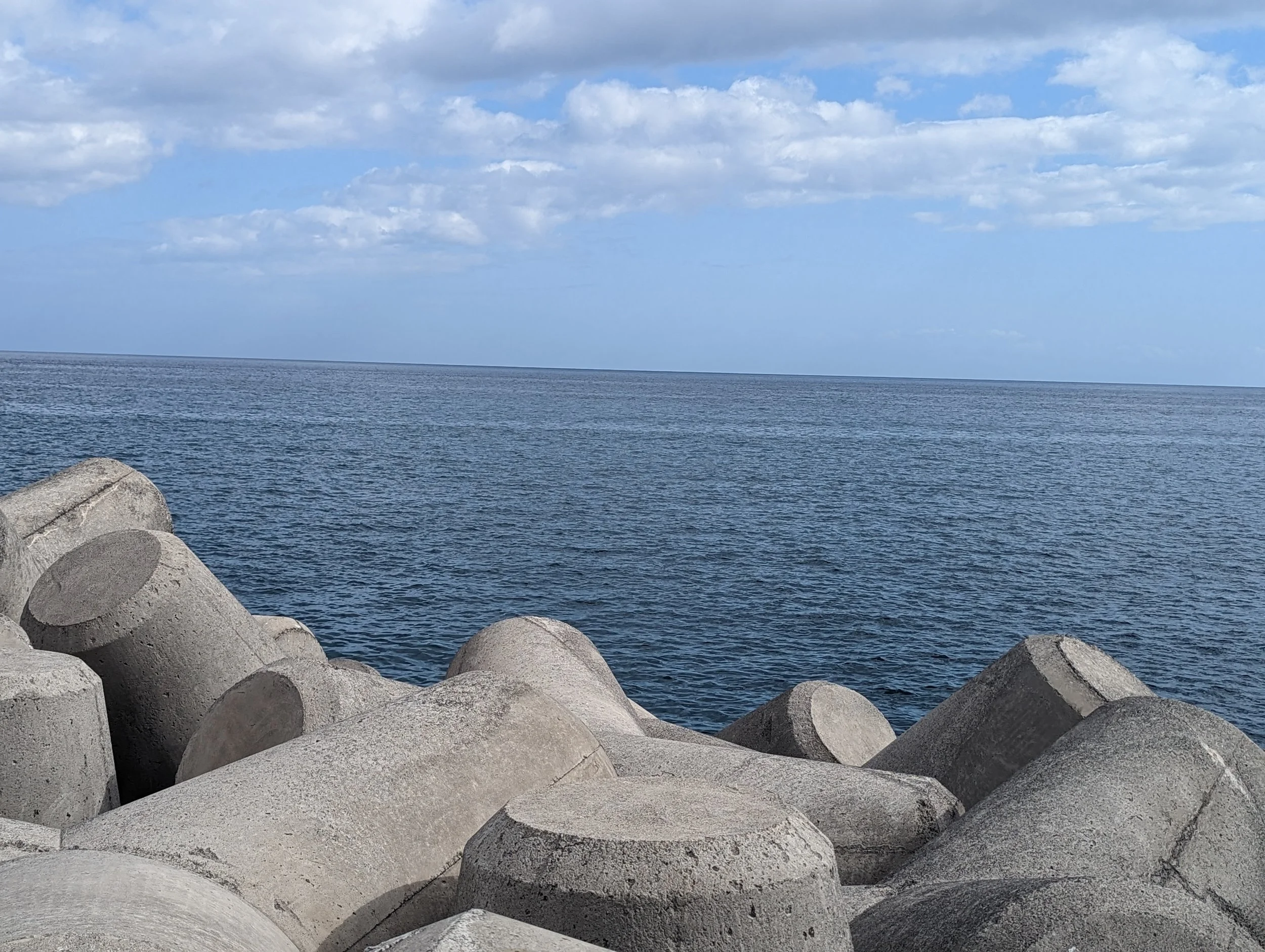 Large concrete tetrapods along a rocky shoreline with a calm ocean and a partly cloudy sky in the background.