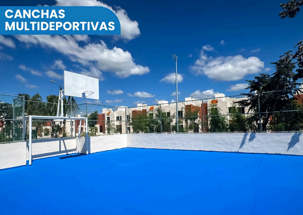 Cancha de deportes multiusos al aire libre con piso azul, rodeada de cerca de malla y con fondo de edificios y árboles, bajo cielo despejado con nubes.