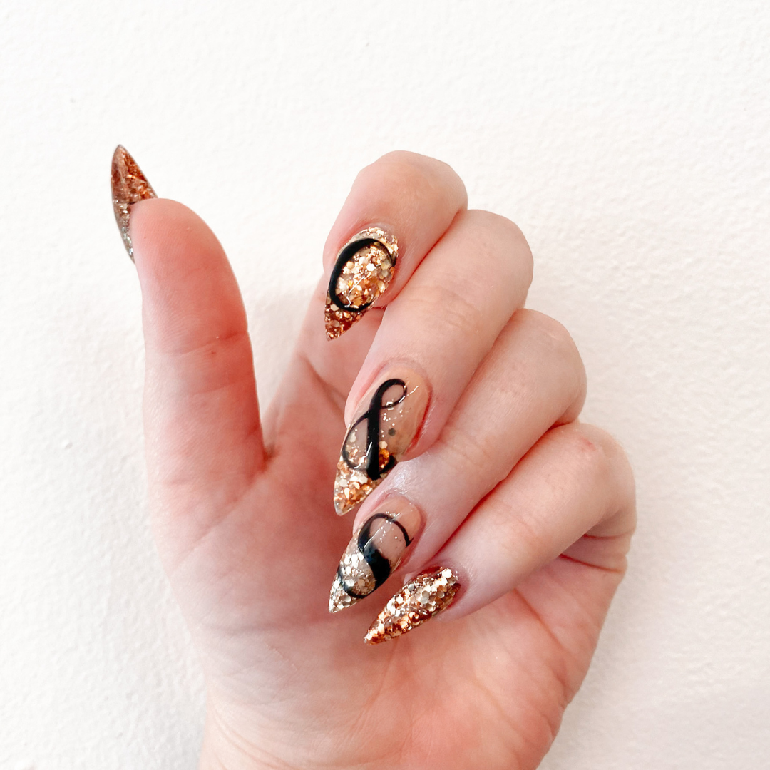 Hand with decorated long almond-shaped nails featuring gold glitter, black accents, and abstract designs.