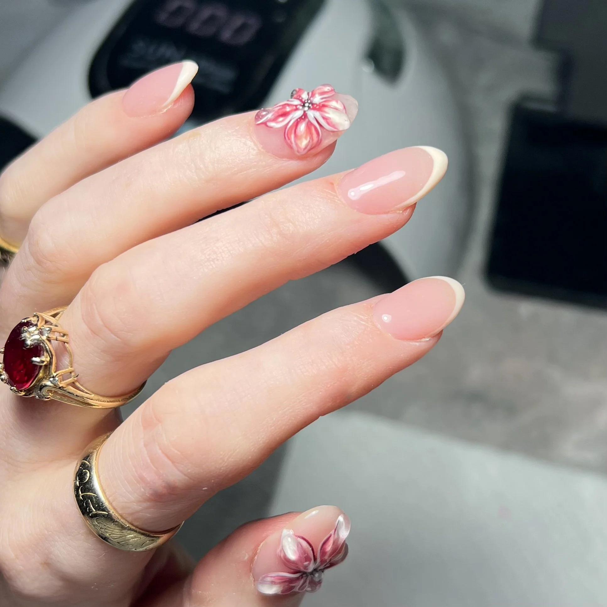 Close-up of a hand with manicured nails, featuring French tip design and 3D pink and white flower nail art on two nails, wearing rings including a gold ring with a red gemstone.