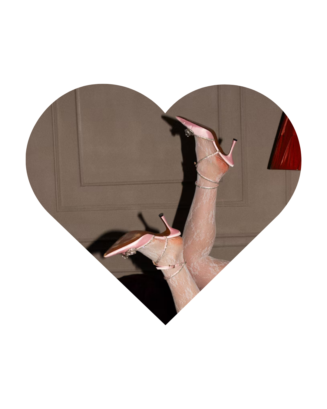 Pair of pink high-heeled shoes with ankle straps, placed on a person's legs wearing lace stockings, against a beige wall and red lampshade.